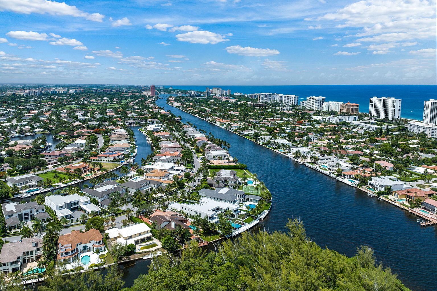 This aerial shot showcases a luxurious waterfront community with stunning homes lining the canals. The properties feature well-manicured lawns, private docks, and swimming pools, creating an idyllic residential setting. The deep blue waterways contrast beautifully with the lush greenery and the clear blue sky, emphasizing the upscale and desirable nature of the neighborhood.