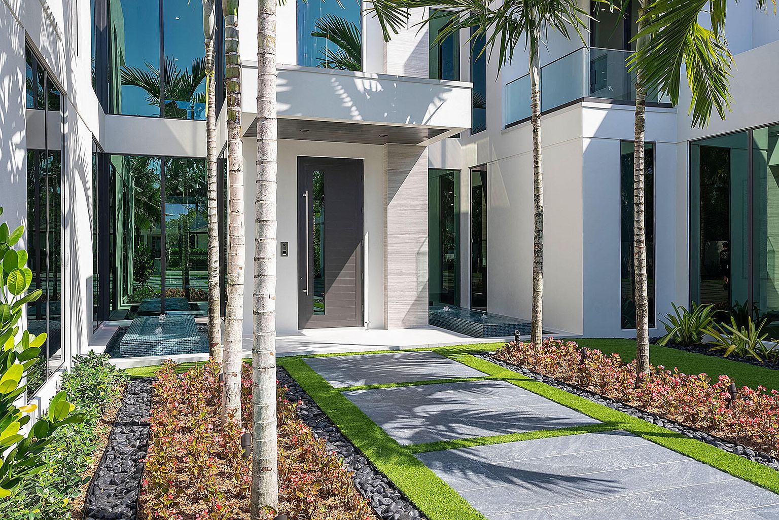 This image showcases the grand entryway of a modern home, featuring a sleek, dark-colored front door with a vertical window pane and a long, silver handle. The walkway is paved with large gray tiles interspersed with strips of artificial turf, leading through a meticulously landscaped garden with black pebbles and vibrant plants. Tall, slender palm trees frame the entrance, adding a touch of tropical elegance to the contemporary design.