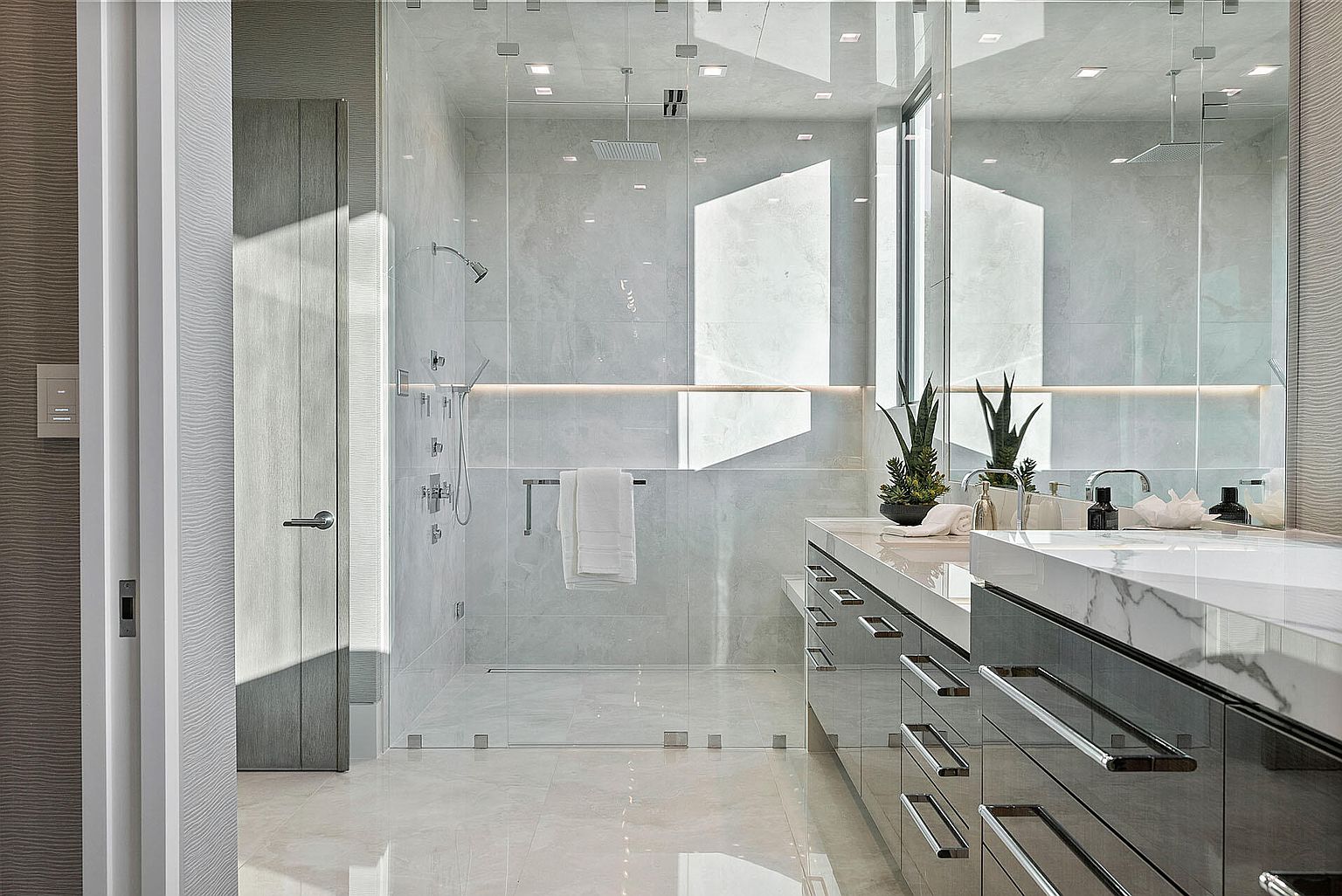 This is a luxurious primary bathroom featuring a glass-enclosed shower with multiple shower heads and a sleek vanity with dual sinks and marble countertops. The bathroom is illuminated by recessed lighting and natural light, creating a bright and airy atmosphere. The overall design is modern and sophisticated, emphasizing clean lines and high-end finishes.