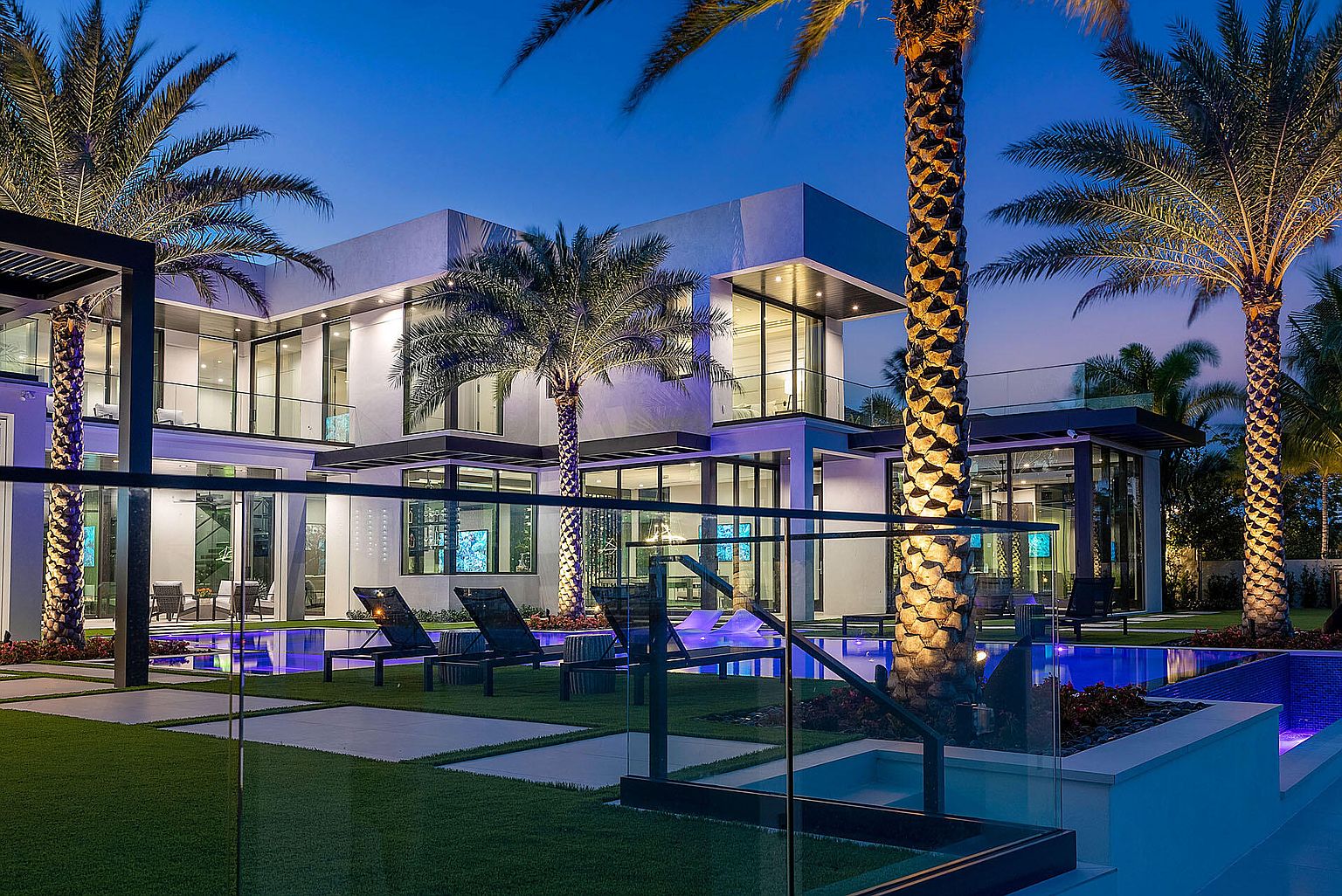 This image showcases the rear exterior of a modern luxury home, emphasizing the pool and outdoor living space. The pool is illuminated with blue lighting, complemented by lounge chairs and lush landscaping. The architecture features clean lines, large windows, and a seamless integration of indoor and outdoor spaces, creating an inviting and sophisticated atmosphere.
