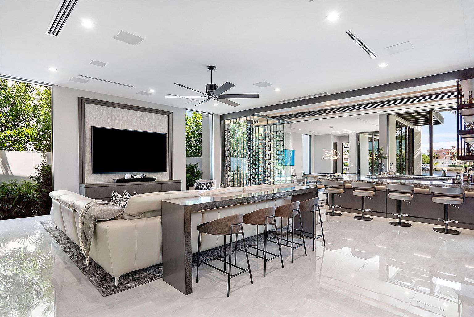 This is a luxurious living room featuring a large television mounted on a textured wall, a modern sofa, and a bar area with stools. A wine rack separates the living area from the bar, and large windows provide ample natural light. The room has a sleek, contemporary design with high-end finishes and a spacious feel.