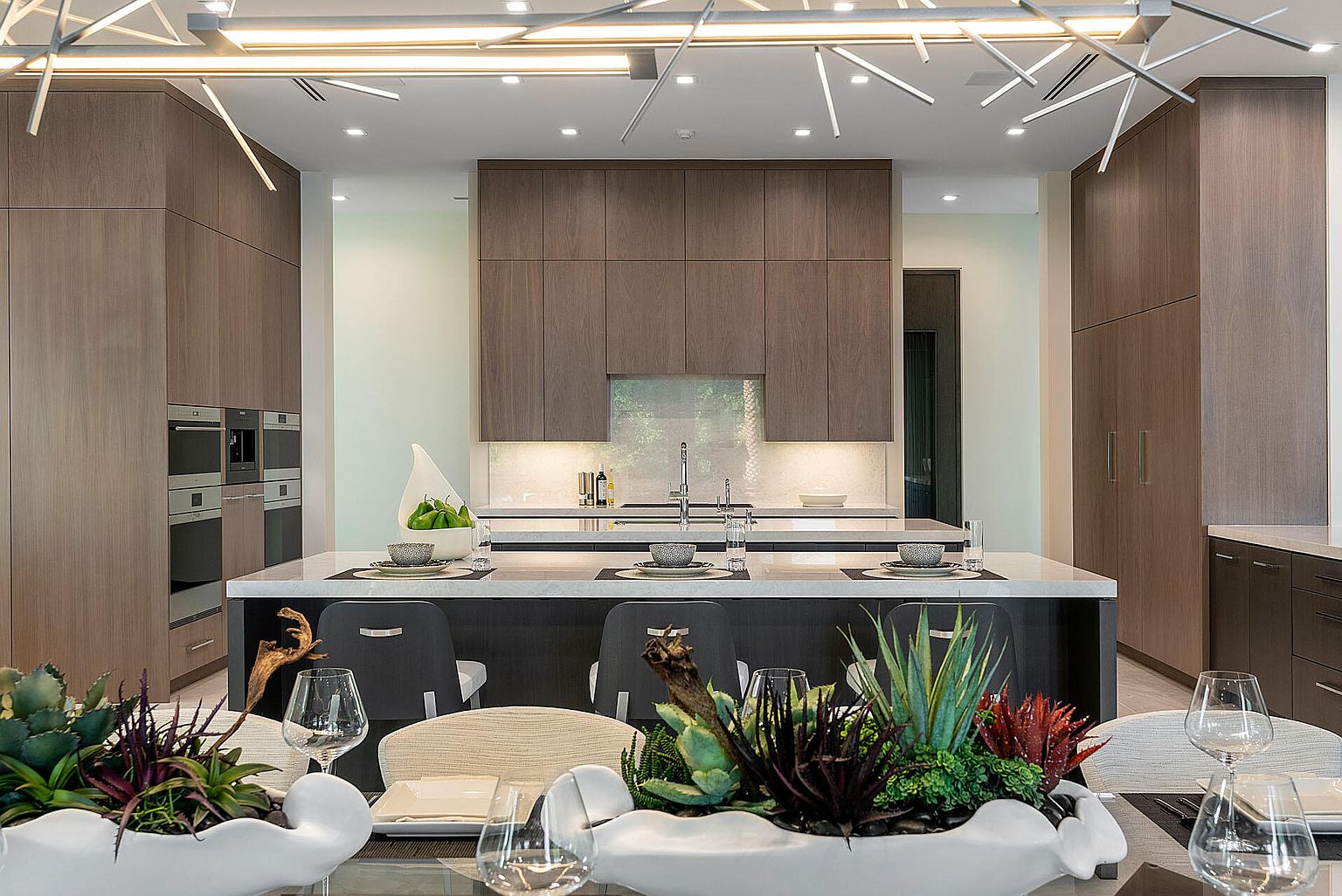 This is a beautifully designed modern kitchen featuring sleek, dark wood cabinetry and a large island with a white countertop. The kitchen is well-lit with modern lighting fixtures, and the overall design is clean and sophisticated. The foreground shows a dining table set with elegant dishware and decorative plants, suggesting a seamless transition between cooking and dining spaces.
