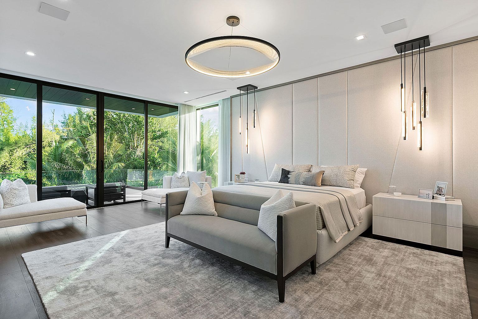 This is a luxurious primary bedroom featuring a large bed with a padded headboard, a gray bench at the foot of the bed, and a modern circular chandelier. Floor-to-ceiling windows offer views of lush greenery, and the room is decorated in a neutral color palette with a large area rug, creating a serene and sophisticated atmosphere. The room also features modern pendant lighting and a sleek nightstand.