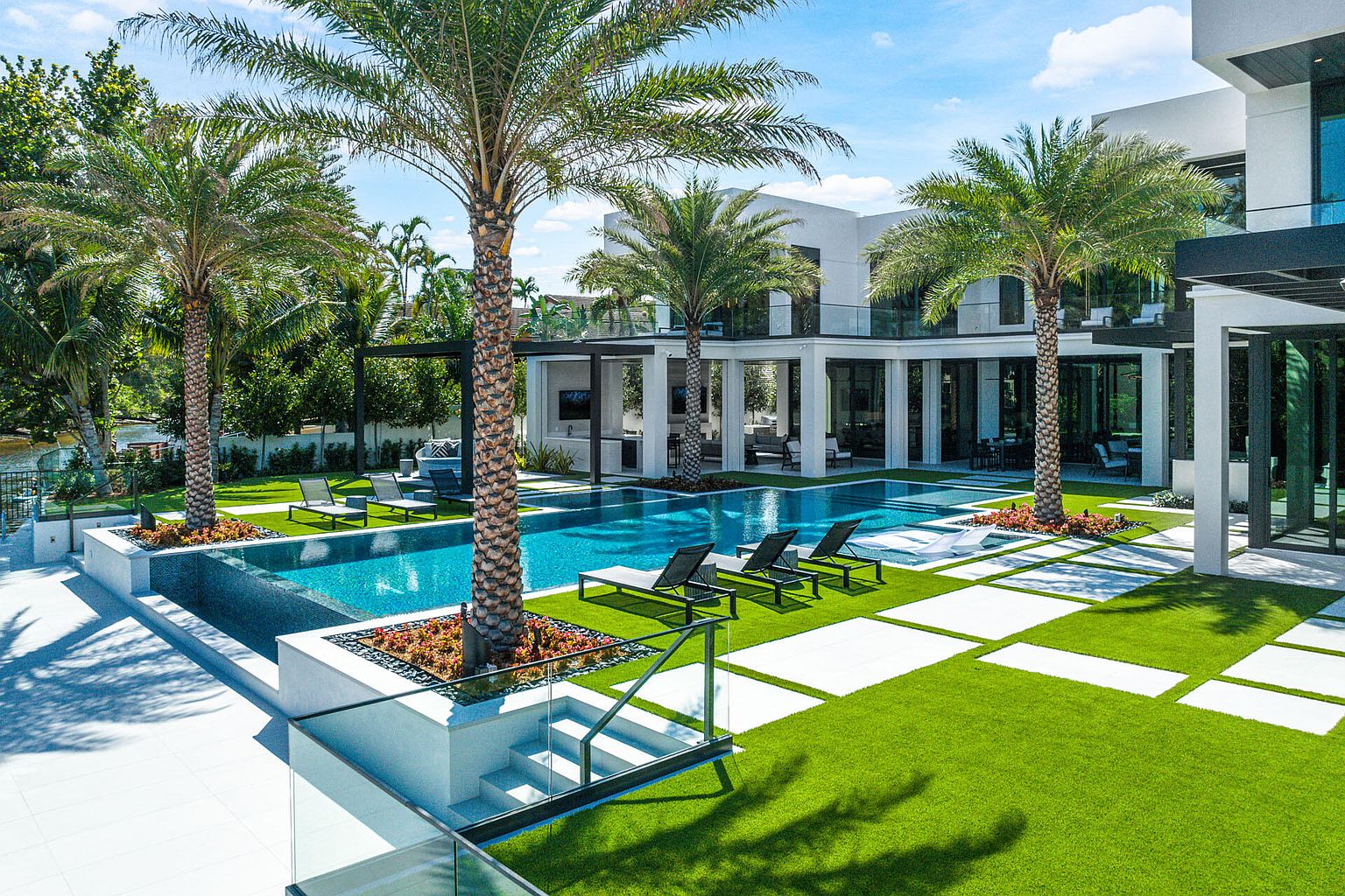 This image showcases a luxurious backyard featuring a modern pool and spa area. The pool is surrounded by lush green turf interspersed with white pavers, creating a visually appealing geometric pattern. Palm trees add a tropical touch, while the contemporary architecture of the house provides a sleek backdrop, suggesting a high-end property perfect for relaxation and entertainment.