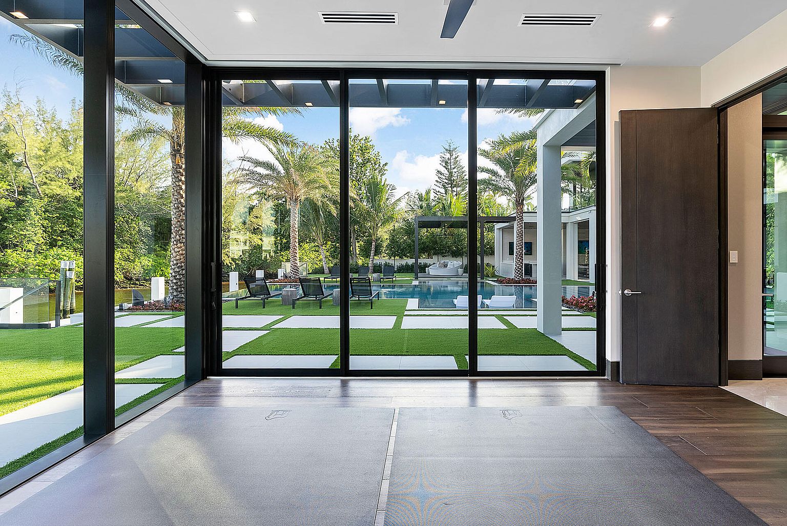 This interior shot showcases a modern living room with a view of a luxurious backyard featuring a pool and manicured lawn. Large sliding glass doors provide ample natural light and create a seamless transition to the outdoor space. The room features sleek flooring and a minimalist design, emphasizing the connection to the outdoor oasis.