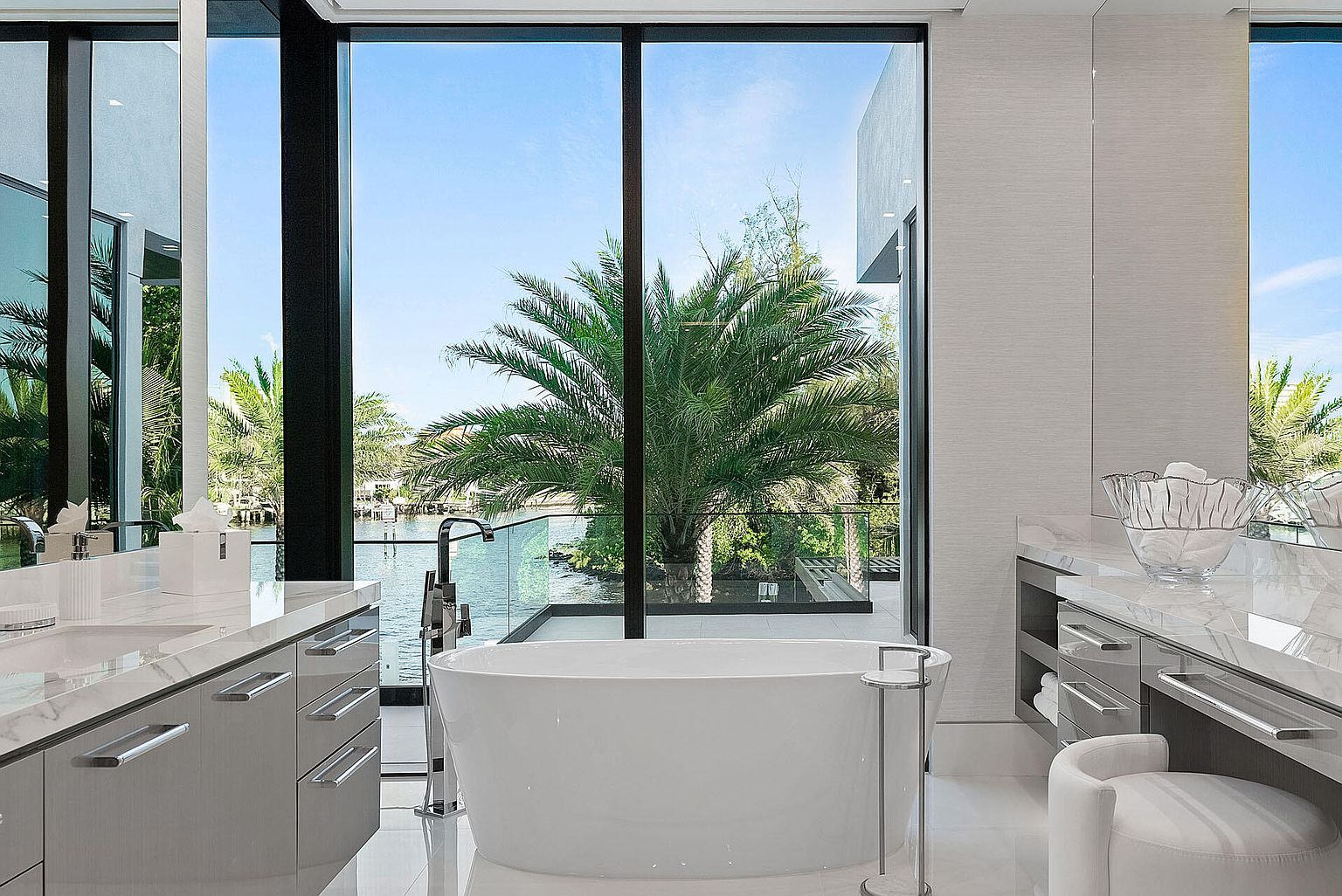 This is a luxurious primary bathroom featuring a freestanding white bathtub positioned in front of a large window with a view of lush greenery and waterfront. The bathroom includes modern vanities with marble countertops and sleek hardware, complemented by a minimalist stool. The overall design is contemporary and spa-like, emphasizing natural light and serene views.