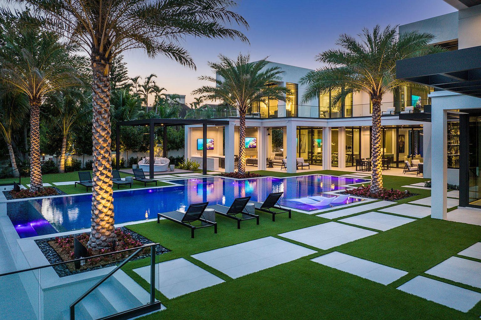 This image showcases a luxurious backyard with a stunning pool area. The modern home features large windows and a sleek design, complemented by palm trees and manicured landscaping. The pool is surrounded by lounge chairs and white tile pathways interspersed with green turf, creating an inviting and upscale outdoor living space.