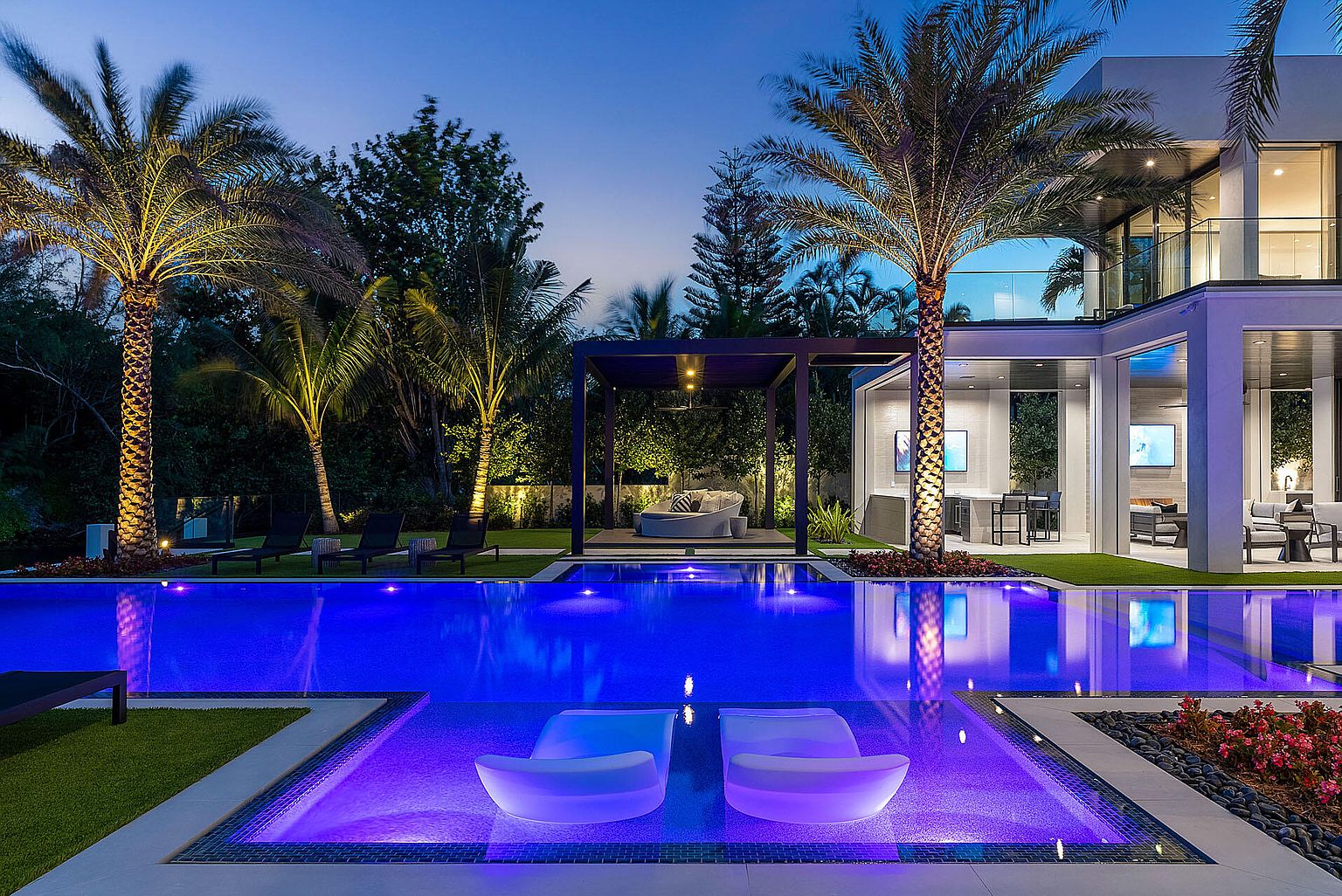 This image showcases a luxurious backyard pool area at dusk, featuring a modern infinity pool with integrated lounge chairs and vibrant blue lighting. The pool is surrounded by lush landscaping, including palm trees, and a contemporary pergola with outdoor seating. The adjacent modern home boasts large windows and sleek architectural lines, creating an inviting and upscale outdoor living space.