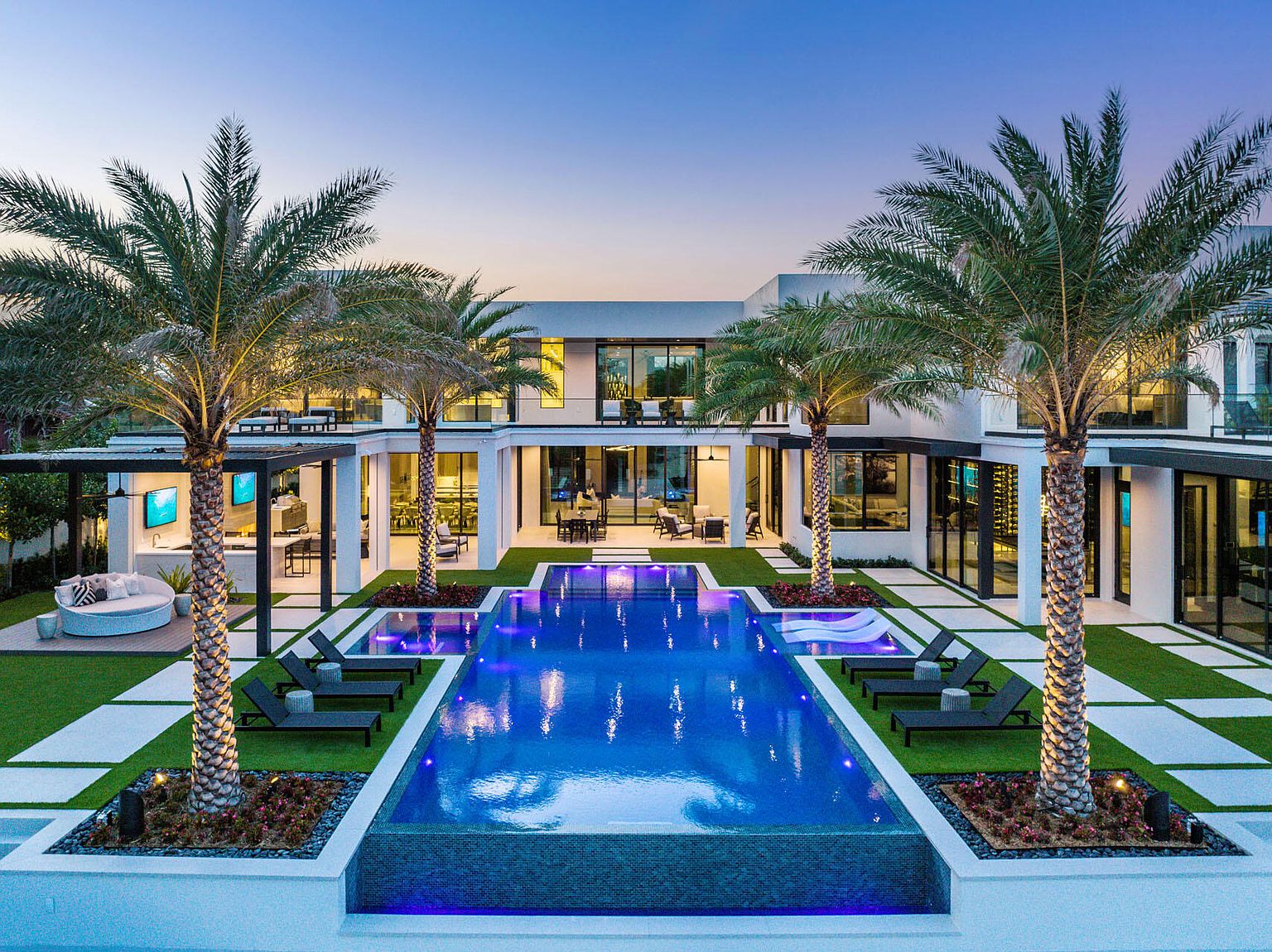 This image showcases a luxurious backyard featuring a rectangular pool with vibrant blue lighting, surrounded by meticulously manicured landscaping. Palm trees frame the view of the modern, white-exterior home with expansive windows and outdoor living spaces. The scene exudes a sense of high-end relaxation and sophisticated outdoor living.