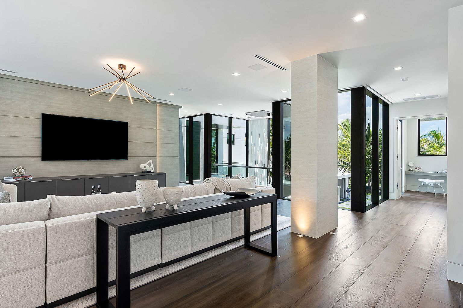 This is a modern living room featuring dark wood flooring and a neutral color palette. A large sectional sofa faces a wall-mounted television above a sleek console. A black console table sits behind the sofa, adding a touch of elegance to the space. Large windows and glass doors provide ample natural light and views of the outdoors.