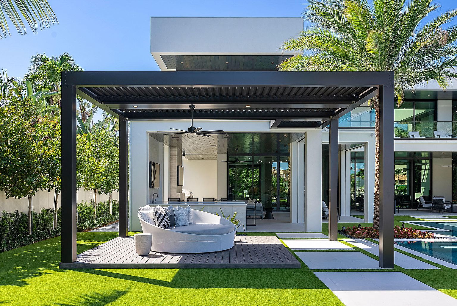 This image showcases a luxurious outdoor patio area with a modern pergola, a stylish round sofa, and a well-maintained artificial lawn. The patio seamlessly connects to the interior through large glass doors, offering an open and airy feel. The design emphasizes clean lines and contemporary aesthetics, perfect for upscale outdoor living.