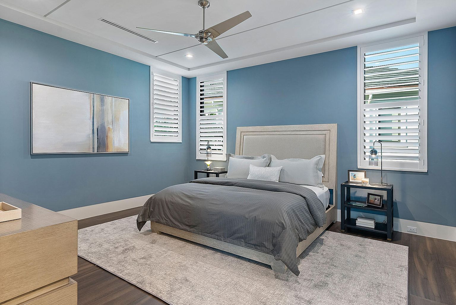 This is a primary bedroom featuring a queen-sized bed with a gray duvet and neutral-toned headboard. The walls are painted a calming blue, complemented by white trim and plantation shutters on the windows. A modern ceiling fan and abstract art add to the room's contemporary style, while a gray rug anchors the space.