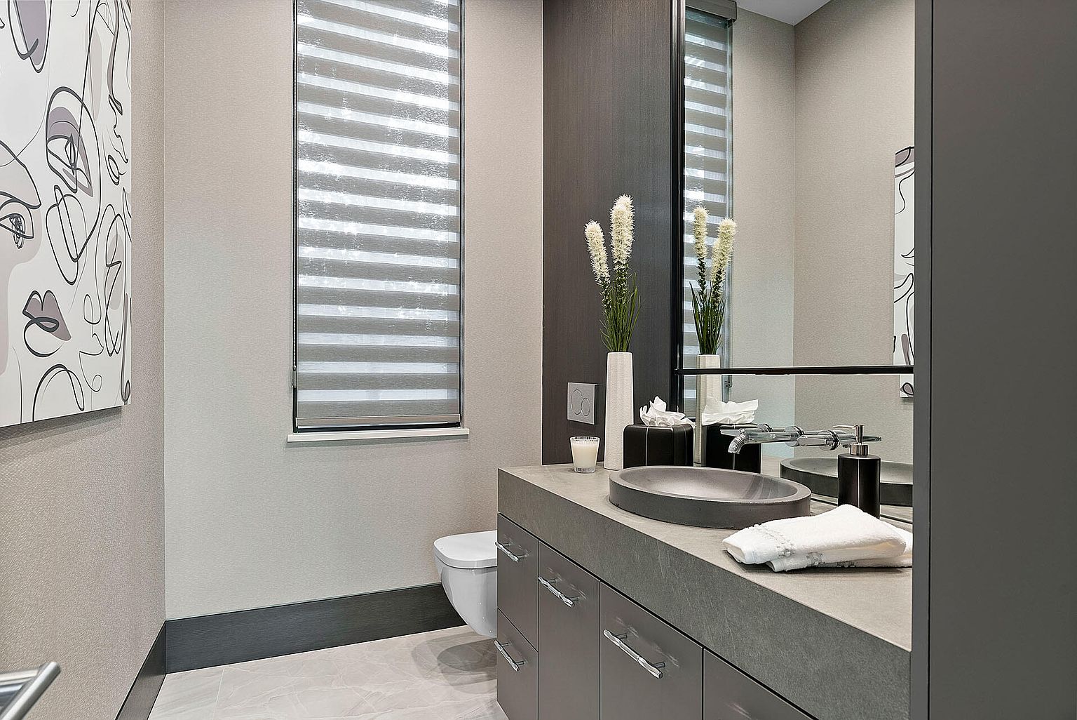 This is a modern guest bathroom featuring a gray vanity with a vessel sink and chrome fixtures. A large mirror reflects the window with horizontal blinds, and a piece of abstract art hangs on the wall. The color palette is neutral, creating a serene and sophisticated atmosphere.
