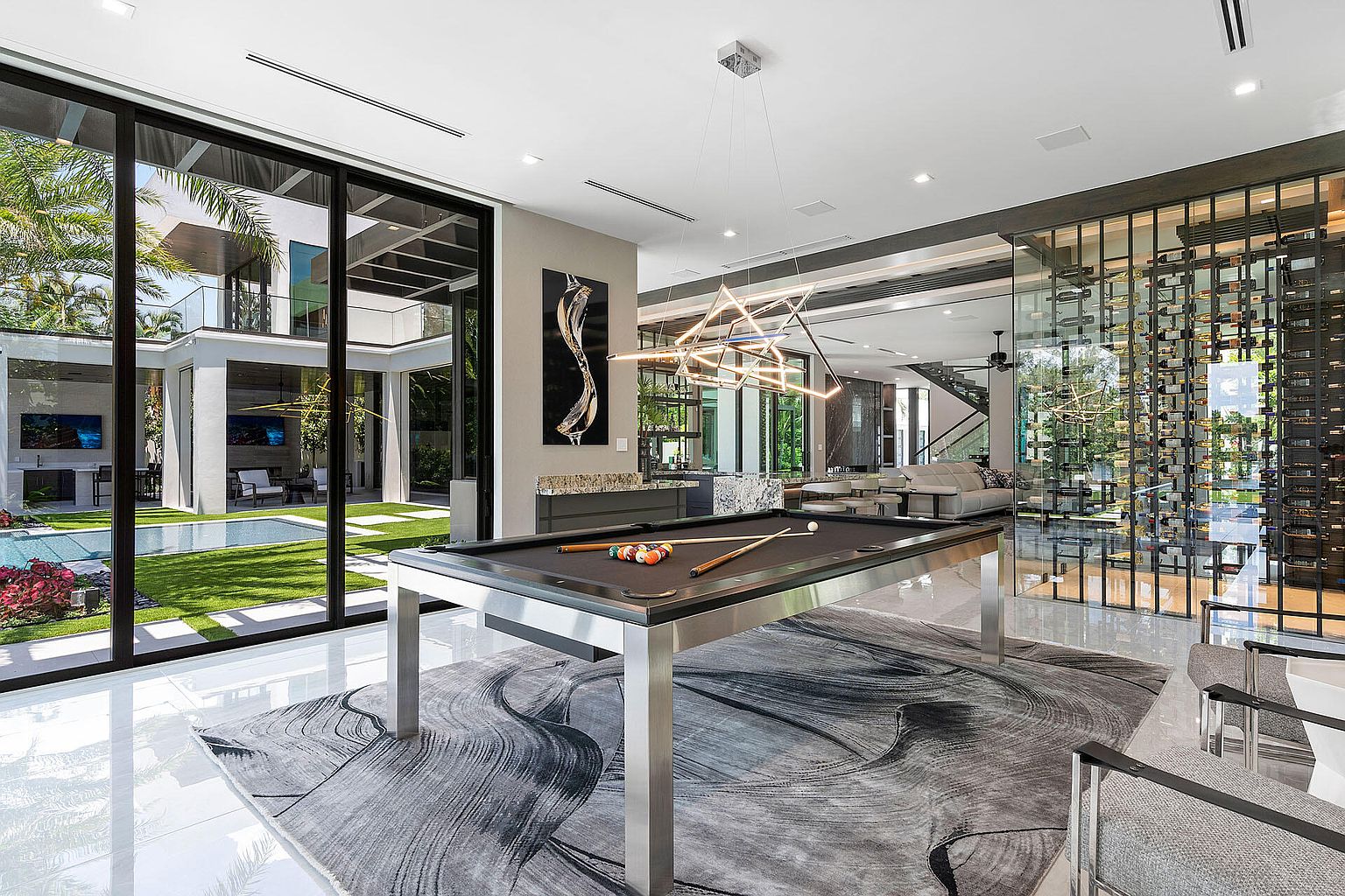 This interior shot showcases a luxurious living room with a modern aesthetic. A pool table sits prominently on a patterned rug, illuminated by a contemporary chandelier. Large windows offer a view of the outdoor pool and landscaping, while a wine cellar adds a touch of sophistication to the space.