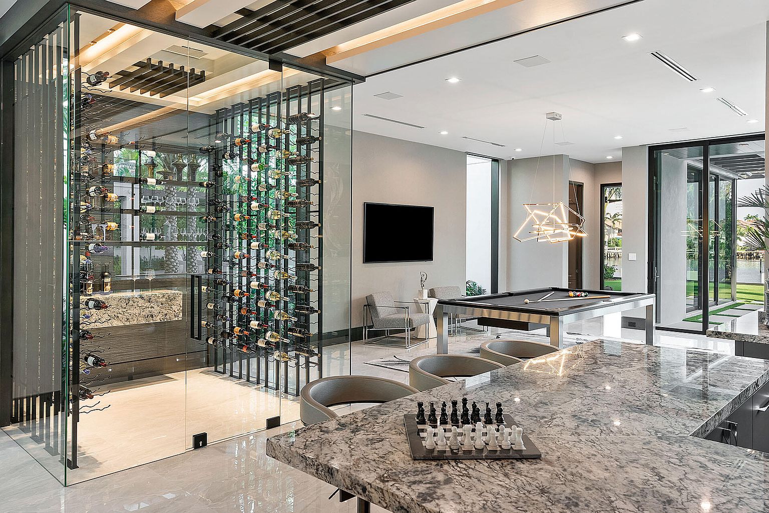 This interior shot showcases a luxurious living space with a modern wine cellar enclosed in glass, a sleek pool table, and a stylish bar area with marble countertops. The room features contemporary lighting fixtures, neutral color palettes, and large windows that offer views of the outdoors, creating an open and sophisticated atmosphere. The perspective is from behind the bar, looking into the living space.
