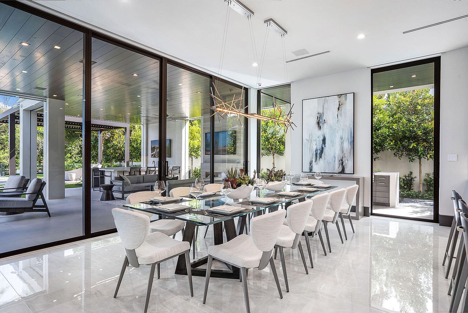 This is a bright and modern dining room featuring a glass-topped dining table with seating for twelve. The room is well-lit with a contemporary chandelier and natural light streaming through large glass doors that offer views of the outdoor space. The polished marble floors and minimalist decor contribute to a luxurious and spacious feel, perfect for entertaining.