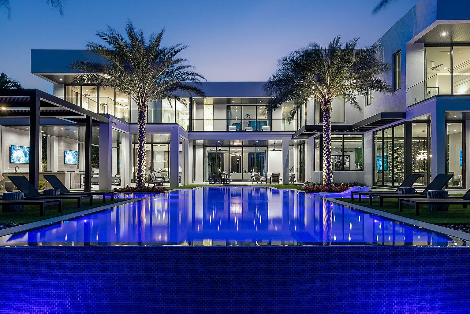 This image showcases a luxurious modern home with a stunning backyard pool at twilight. The pool's blue lighting creates a vibrant reflection of the house, which features large windows and palm trees on either side. Lounge chairs are positioned poolside, suggesting a relaxing outdoor living space.
