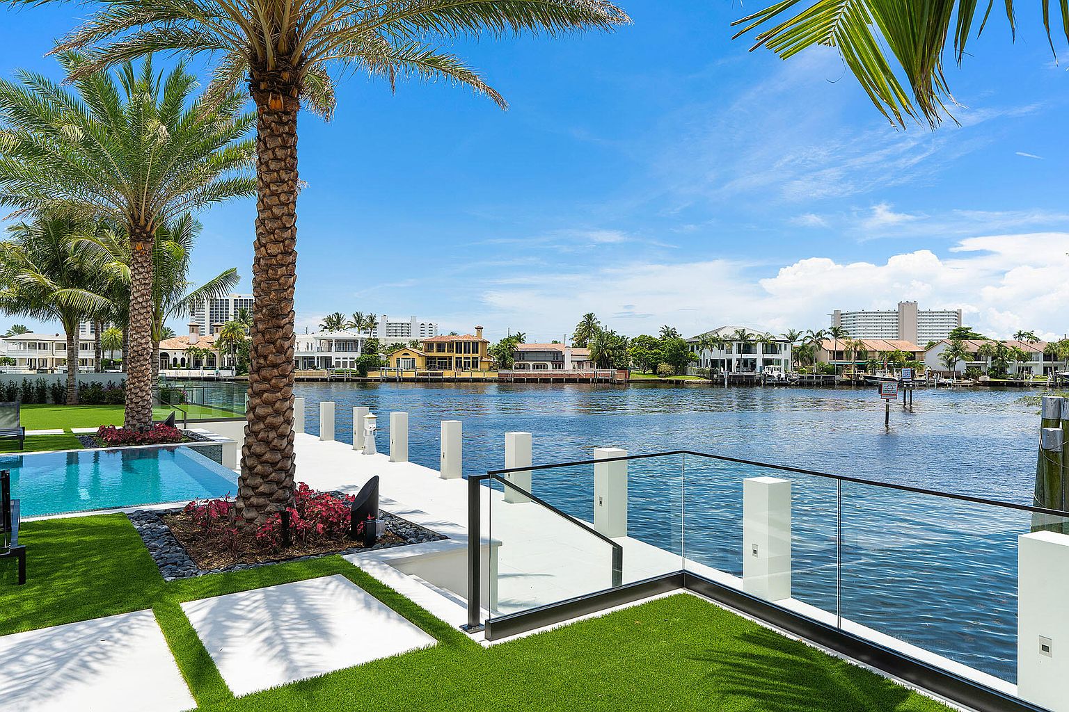 This image showcases a luxurious waterfront property featuring a modern pool and spa area. The design incorporates clean lines, with white pavers and artificial turf creating a sleek, contemporary aesthetic. A glass railing provides unobstructed views of the waterway and surrounding upscale homes, enhancing the property's appeal.