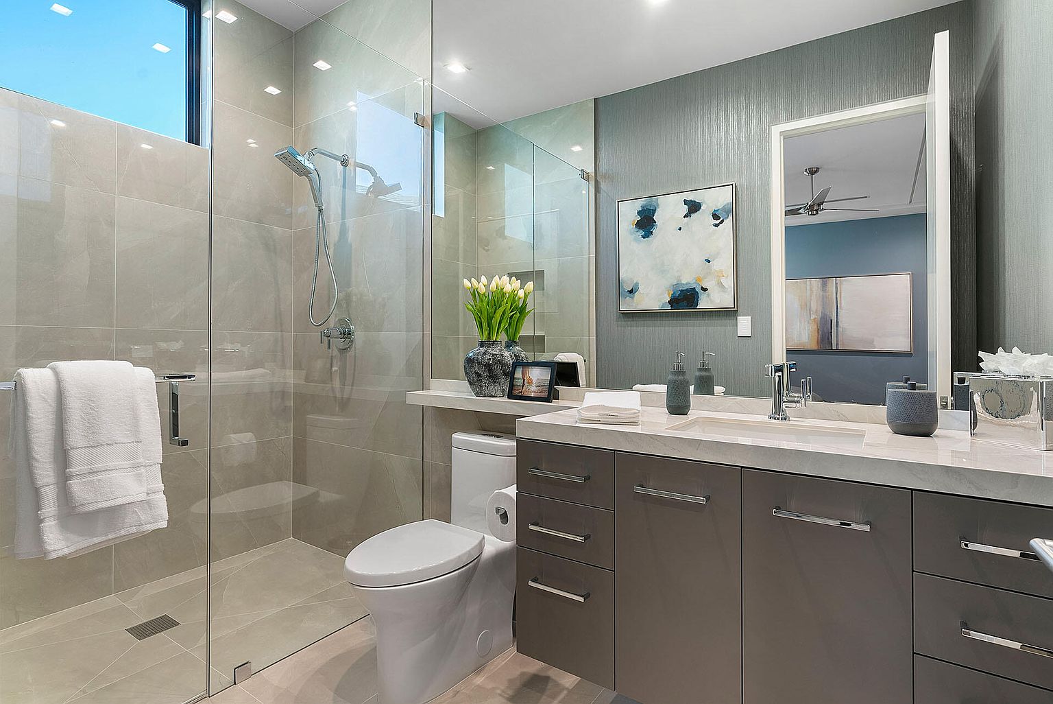This is a modern bathroom featuring a glass-enclosed shower with a rainfall showerhead, a floating vanity with a marble countertop, and sleek gray cabinetry. The walls are painted in a muted blue-gray tone, complemented by an abstract art piece. The overall impression is clean, contemporary, and luxurious.