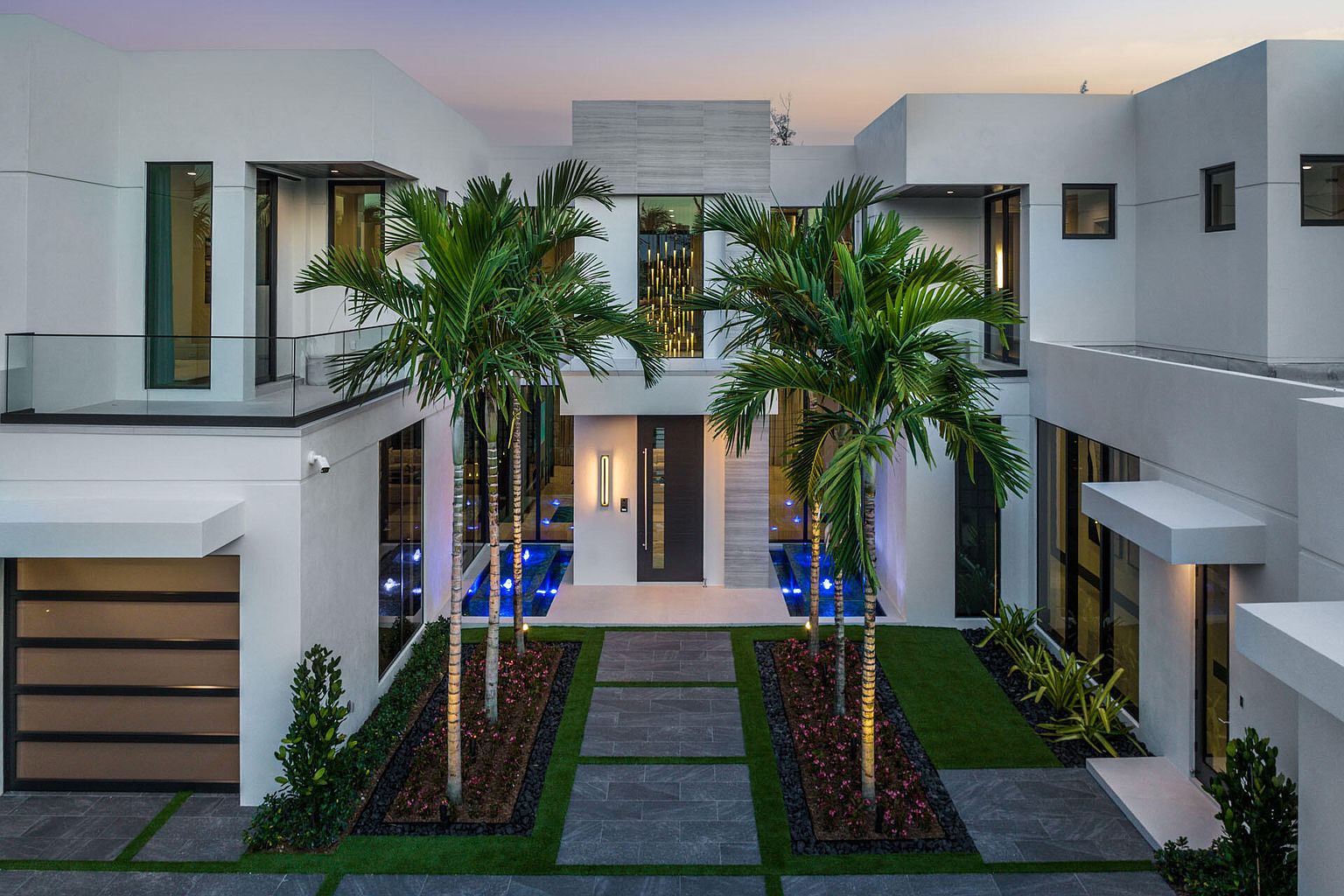 This is a striking front view of a modern, two-story home featuring a clean, white exterior. The entrance is accentuated by two palm trees flanking a walkway of gray stone pavers, leading to a dark-colored front door. The property includes a garage on the left and is landscaped with a combination of green grass, dark gravel, and flowering plants, creating a sophisticated and inviting curb appeal.
