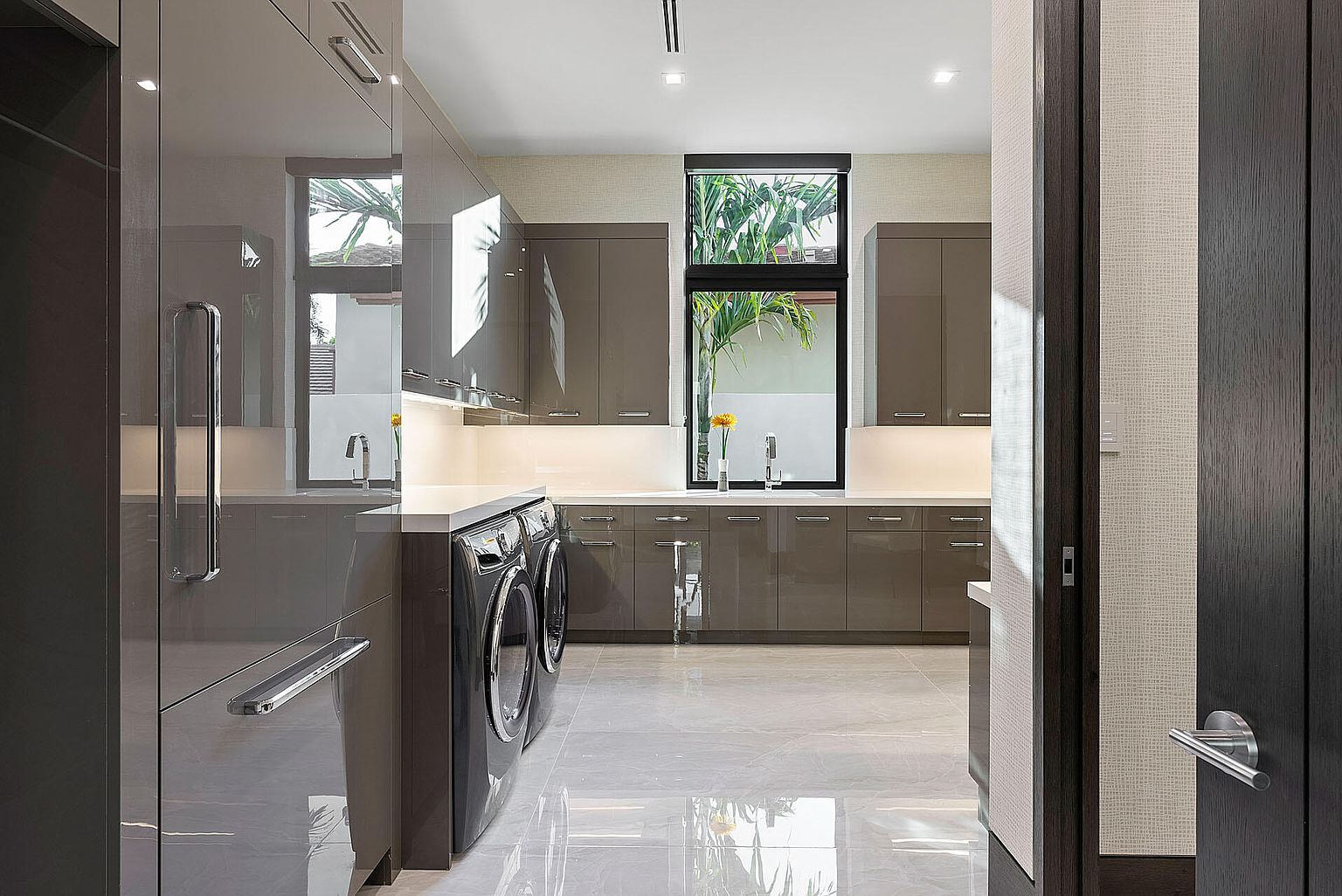 This is a modern laundry room featuring sleek, glossy gray cabinetry and countertops. A front-loading washer and dryer are visible, and a large window provides natural light and a view of greenery. The room has a clean and minimalist aesthetic, with light-colored tile flooring.