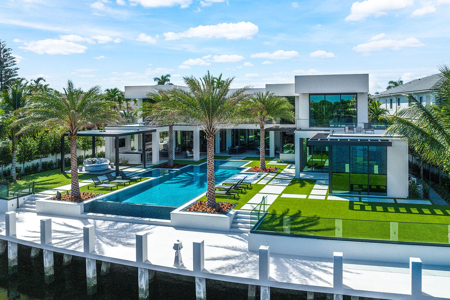 This aerial view showcases a luxurious modern home with a stunning backyard oasis. The property features a sleek, rectangular pool surrounded by palm trees, manicured lawns with geometric stone pathways, and a stylish outdoor lounge area. A private dock extends into the water, adding to the property's waterfront appeal.