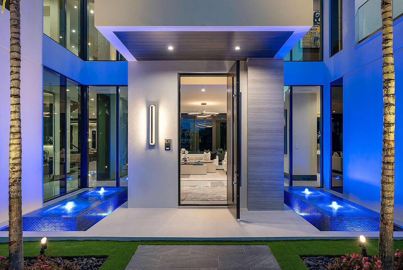 This is a striking entryway to a modern home, featuring a grand pivot door flanked by water features with blue-lit fountains. The exterior is a blend of smooth white walls and textured gray panels, illuminated by sleek vertical light fixtures. Palm trees frame the entrance, adding a touch of tropical elegance.
