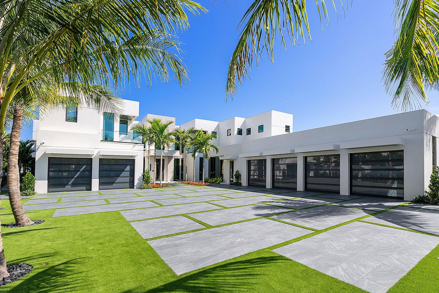 This is a front exterior view of a modern, multi-story home featuring a white facade, multiple garage doors with dark frames, and a meticulously designed driveway with grass accents. Palm trees frame the house, adding a tropical touch to the property. The clear blue sky enhances the bright and clean aesthetic of the home, suggesting a luxurious and well-maintained property.