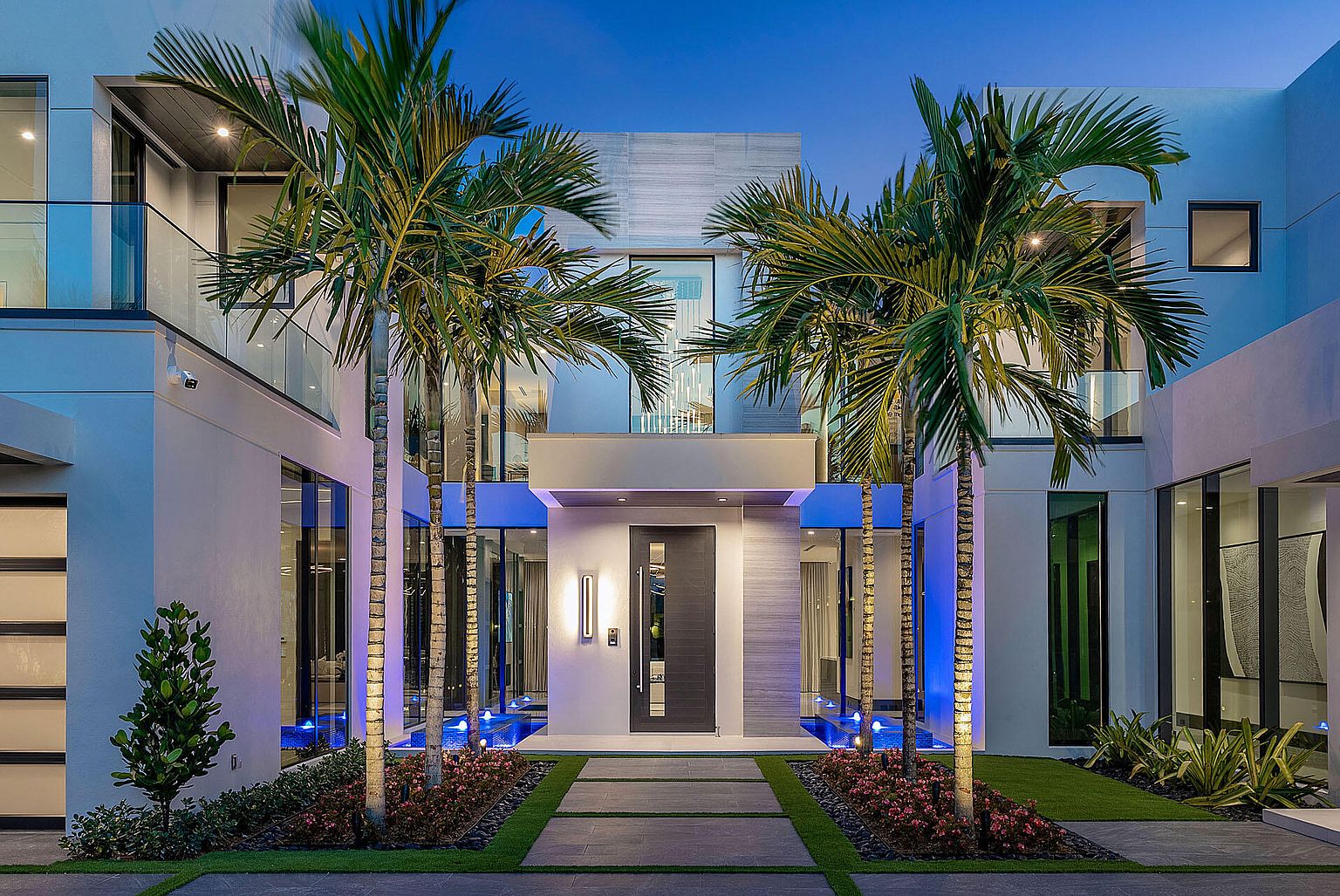 This is a striking front view of a modern home's entryway at dusk, featuring a sleek, dark front door flanked by illuminated palm trees and water features. The architectural design emphasizes clean lines and a minimalist aesthetic, with large windows and a combination of white and gray exterior finishes. The well-manicured landscaping and subtle blue lighting create an inviting and luxurious ambiance.