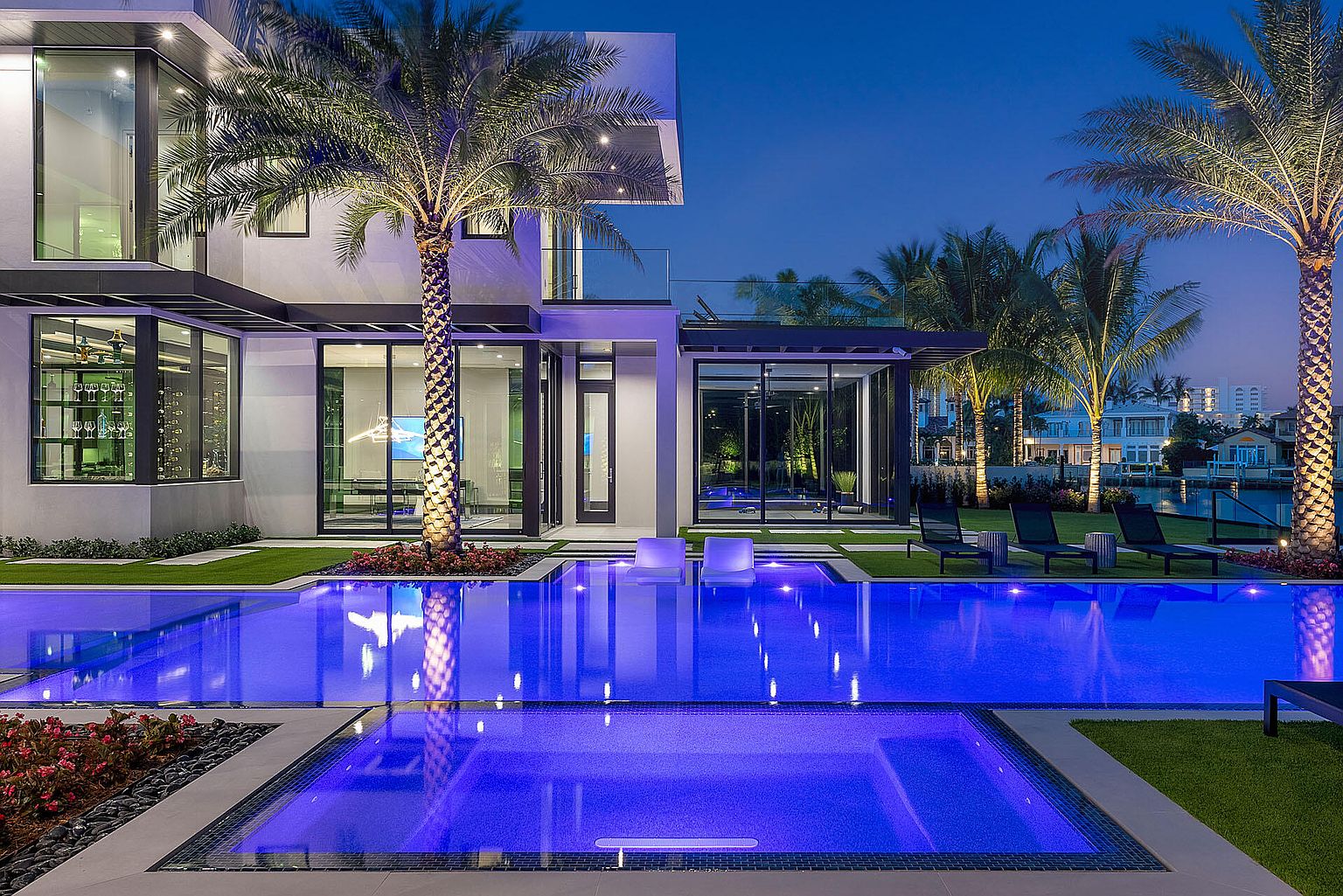 This image showcases a luxurious modern home with a stunning pool and spa area. The pool features vibrant blue lighting, creating a captivating ambiance, while palm trees add a tropical touch. The architecture is sleek and contemporary, with large glass windows offering views into the home's interior, and the overall impression is one of high-end living and relaxation.