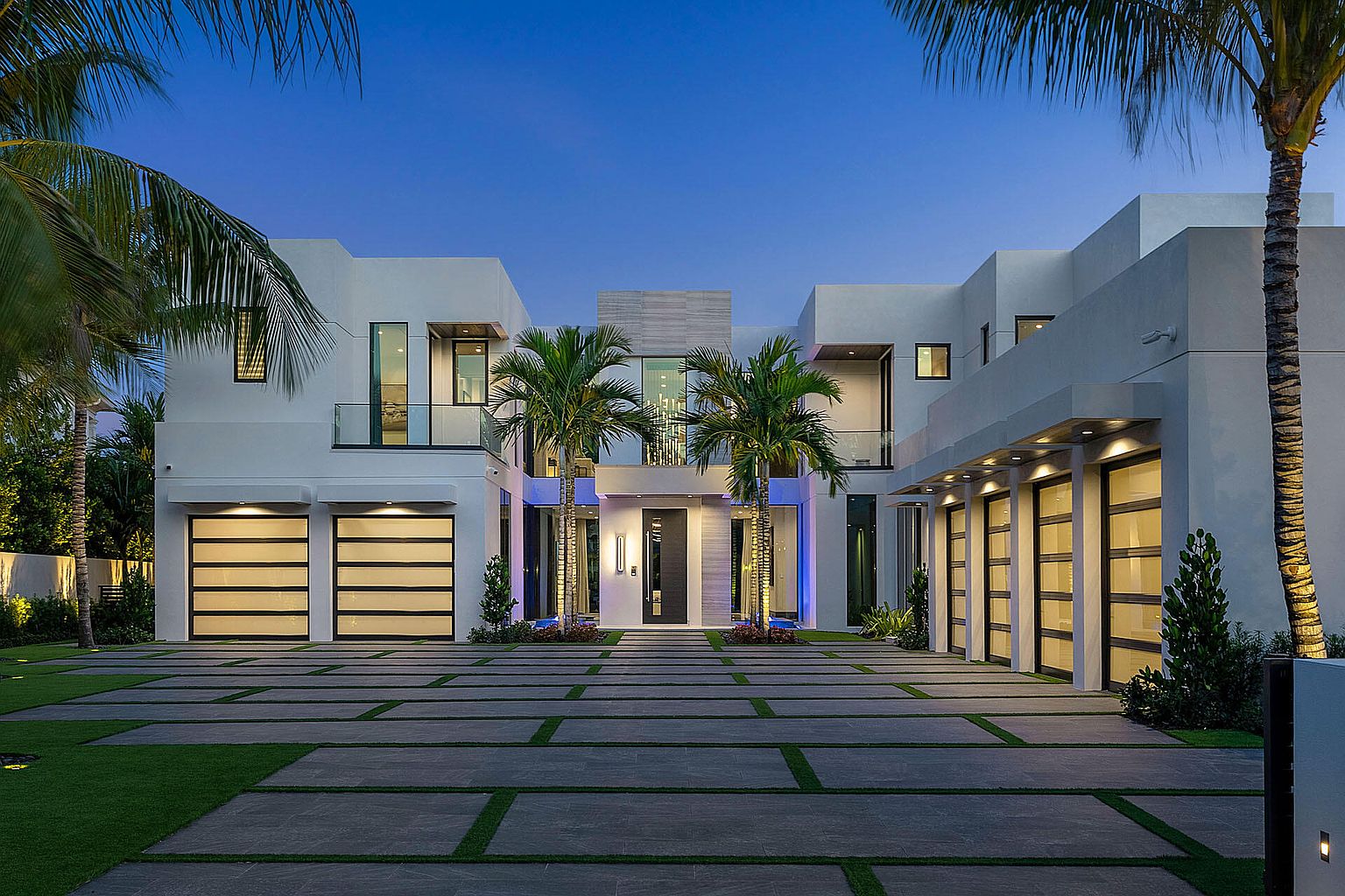 This is a striking front view of a modern, two-story home featuring a symmetrical design with clean lines and a white exterior. The property boasts multiple garage doors with glass panels, lush landscaping including palm trees, and a paved driveway with grass accents. The overall impression is one of luxury and contemporary elegance, perfect for a high-end real estate listing.