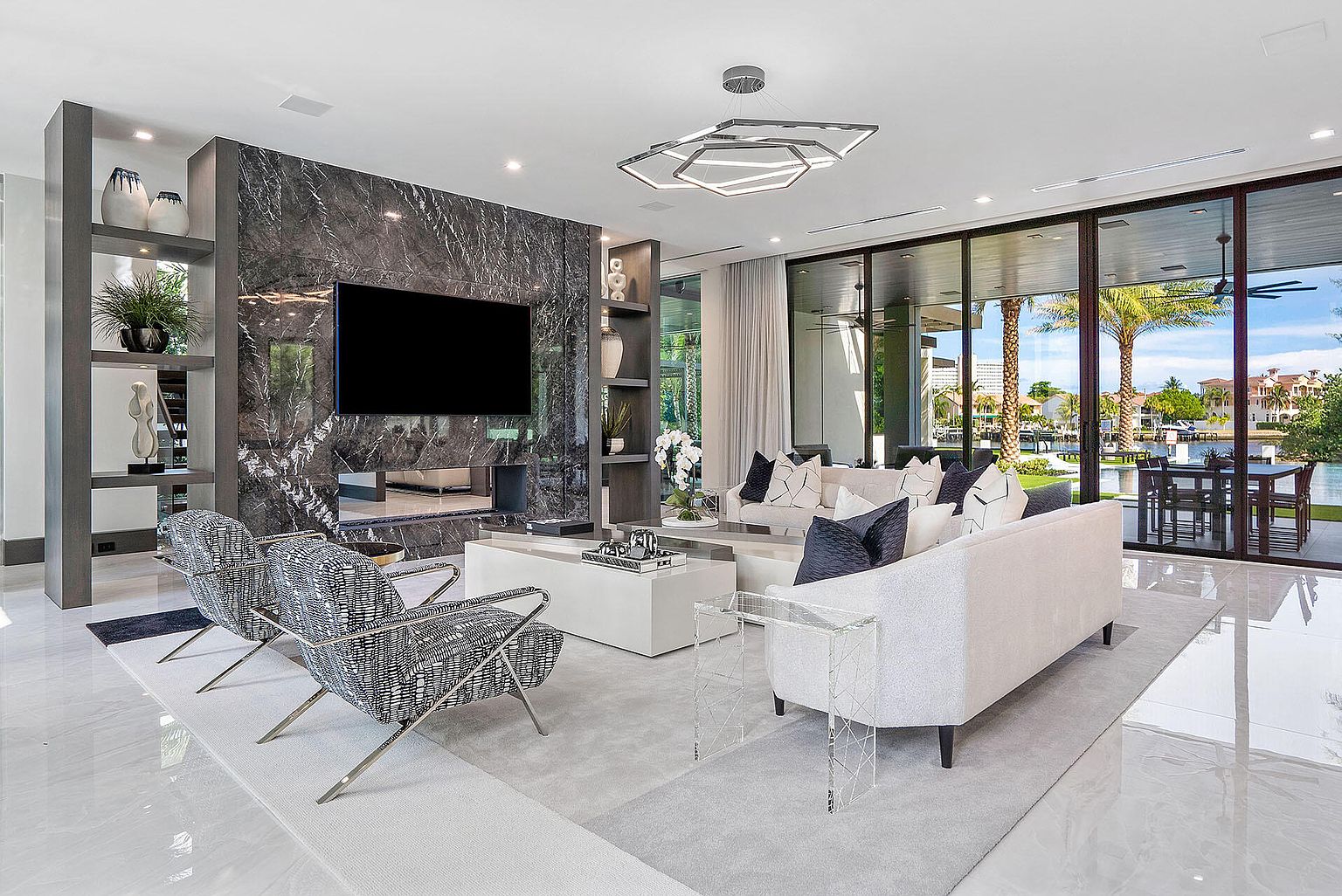 This is a luxurious living room featuring a marble accent wall with a large flat-screen TV and built-in shelving. The room is furnished with modern seating, including two patterned chairs and a white sofa with decorative pillows. Large windows offer a view of the waterfront, and a unique geometric chandelier illuminates the space, creating an atmosphere of sophisticated elegance.