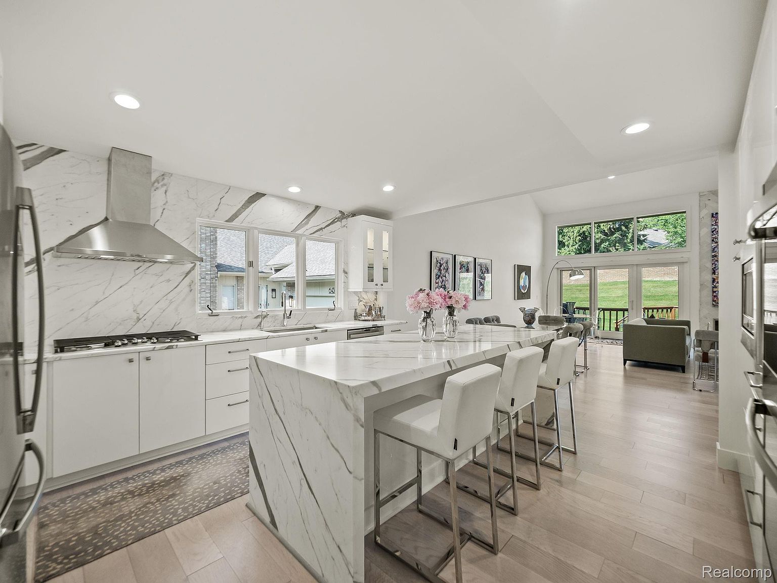 This is a bright, open-concept kitchen featuring white marble countertops and backsplash, stainless steel appliances, and a large island with bar seating. The kitchen seamlessly flows into the living area, creating an inviting space for entertaining. Large windows provide ample natural light and views of the outdoors.