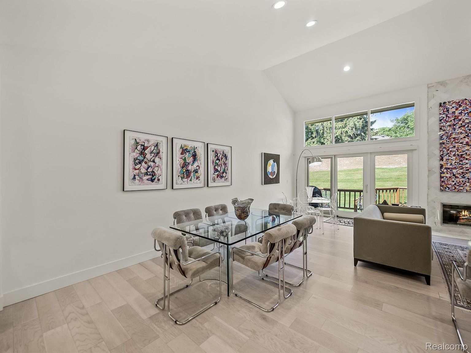 This interior shot showcases a bright and airy dining room with a modern aesthetic. The room features a glass-top dining table surrounded by upholstered chairs with chrome frames, complemented by abstract artwork on the wall. Large windows and sliding glass doors provide ample natural light and a view of the outdoor space, enhancing the room's open and inviting atmosphere.