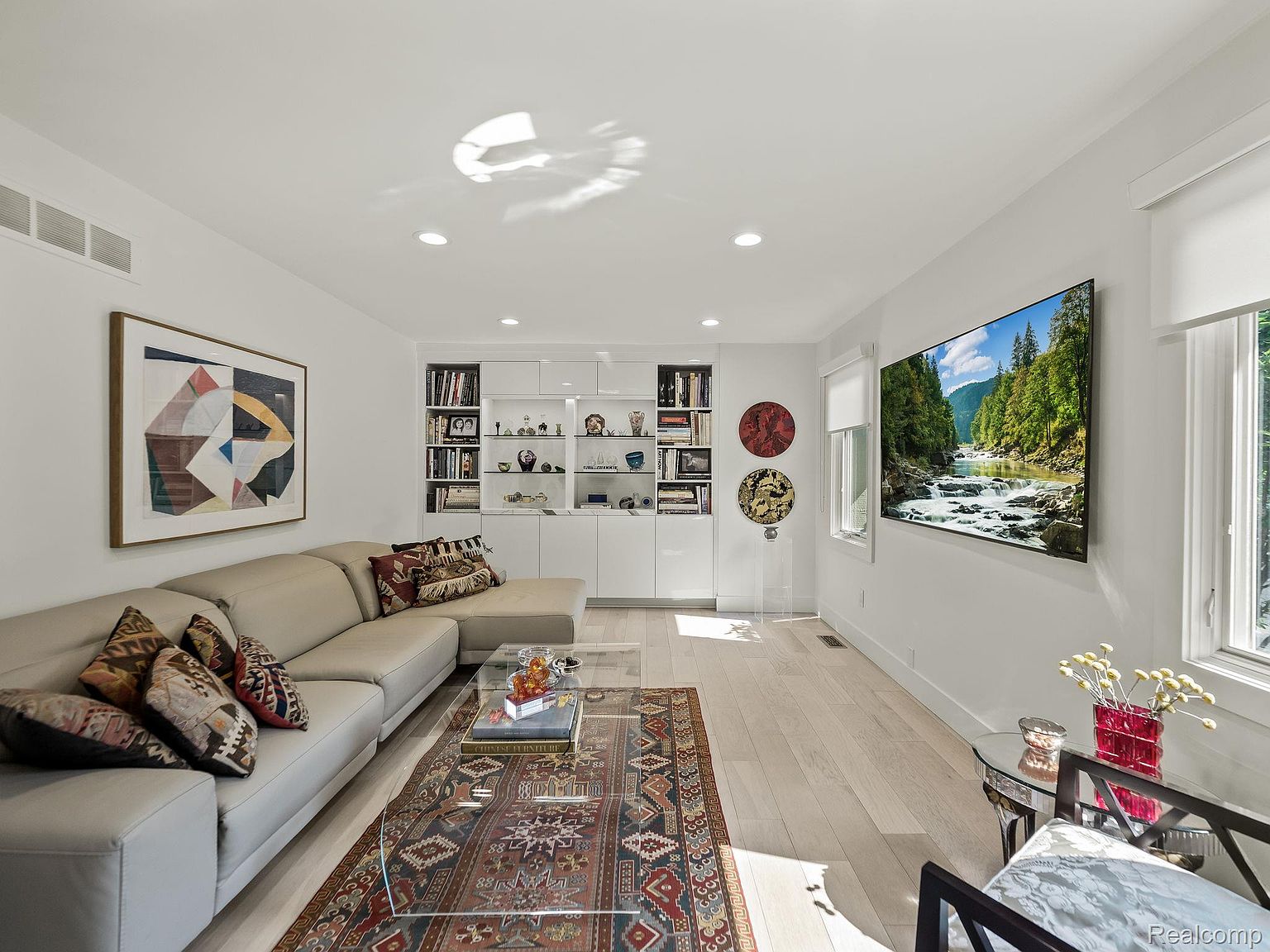 This is an interior shot of a living room featuring a large sectional sofa with decorative pillows, a glass coffee table on top of an ornate rug, and a built-in shelving unit. A large television displaying a nature scene is mounted on the wall, and artwork hangs above the sofa. The room is bright and airy with light wood flooring, creating a modern and inviting atmosphere.