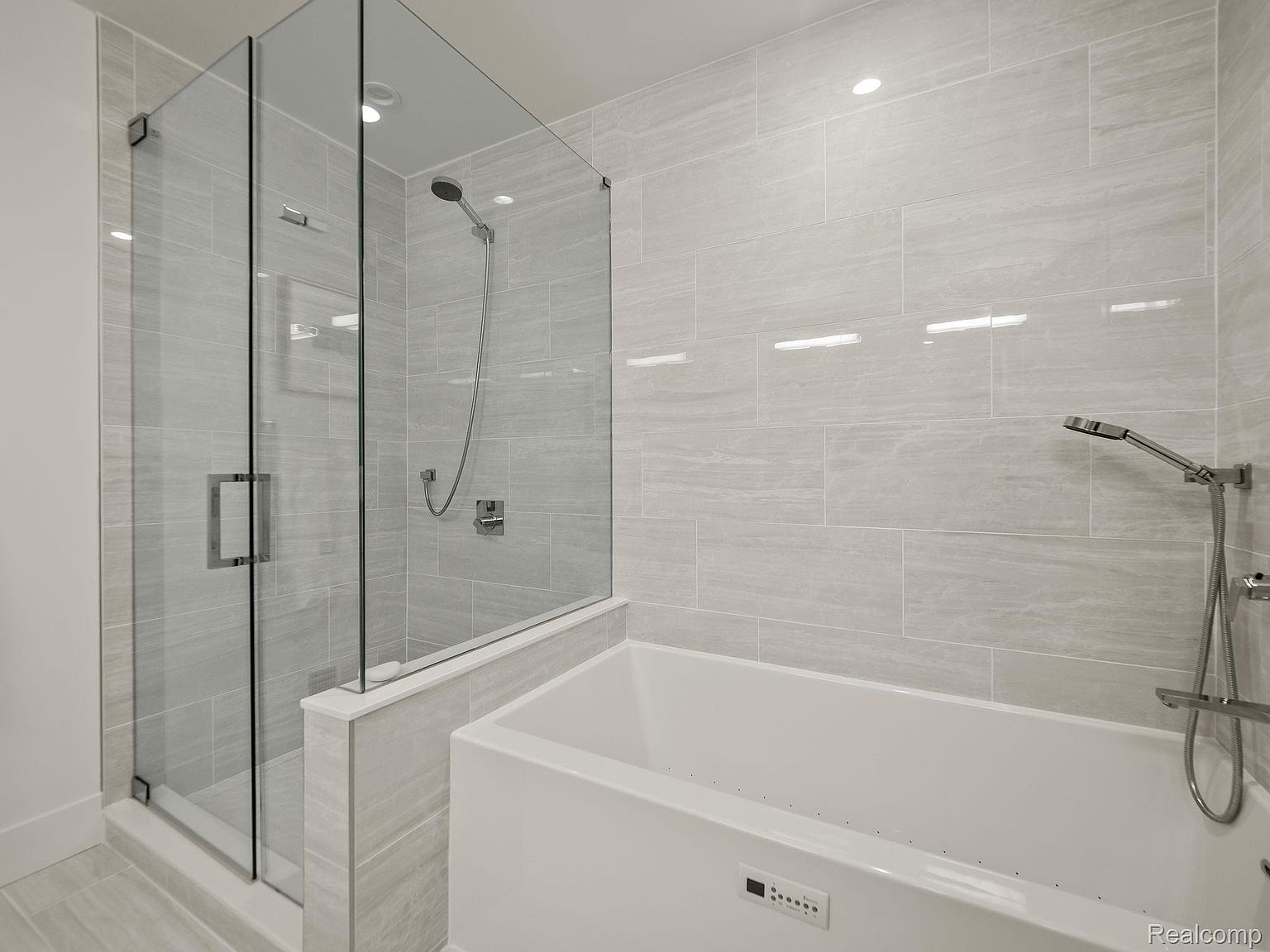 This is a modern primary bathroom featuring a glass-enclosed shower and a soaking tub. The walls are tiled in a light gray, horizontally laid pattern, creating a sleek and contemporary look. The shower has a rainfall showerhead and a handheld sprayer, while the tub has jets and integrated controls.