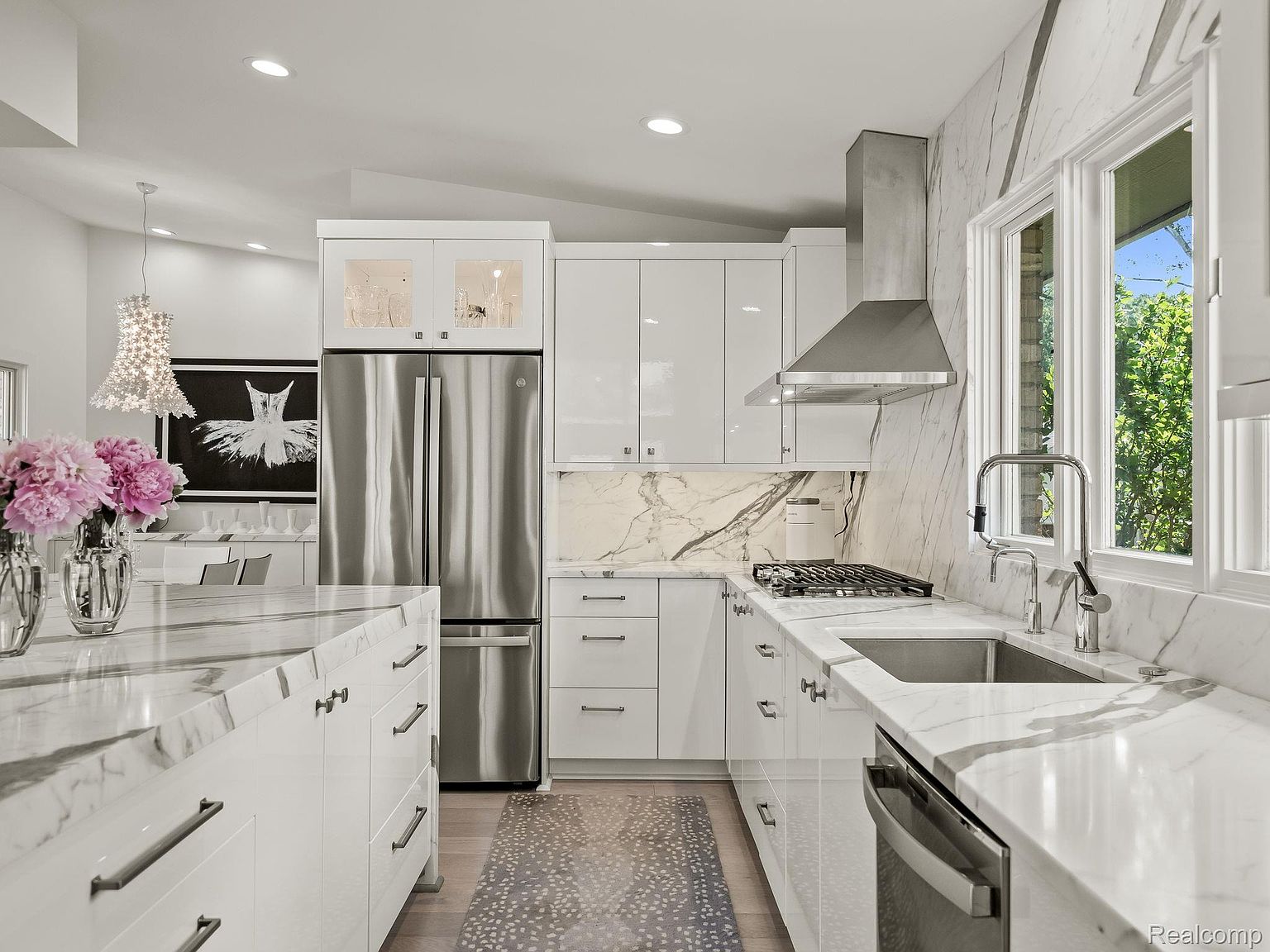 This is a bright, modern kitchen featuring white cabinetry, stainless steel appliances, and marble countertops. The kitchen is well-lit with recessed lighting and natural light from a window overlooking greenery. The overall impression is clean, luxurious, and functional, perfect for a modern home.