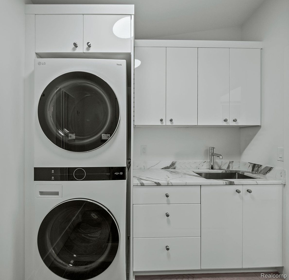 This is an interior shot of a laundry room featuring a stacked LG washer and dryer set. The room also includes white cabinetry with silver hardware, a marble countertop, and a stainless steel sink. The overall impression is clean and modern.