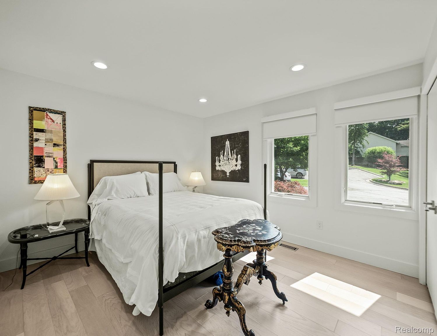 This is a bright and airy primary bedroom featuring a four-poster bed with white linens, hardwood floors, and recessed lighting. The room is decorated with modern art and stylish side tables, creating a serene and inviting atmosphere. Natural light streams in through the windows, enhancing the room's appeal.