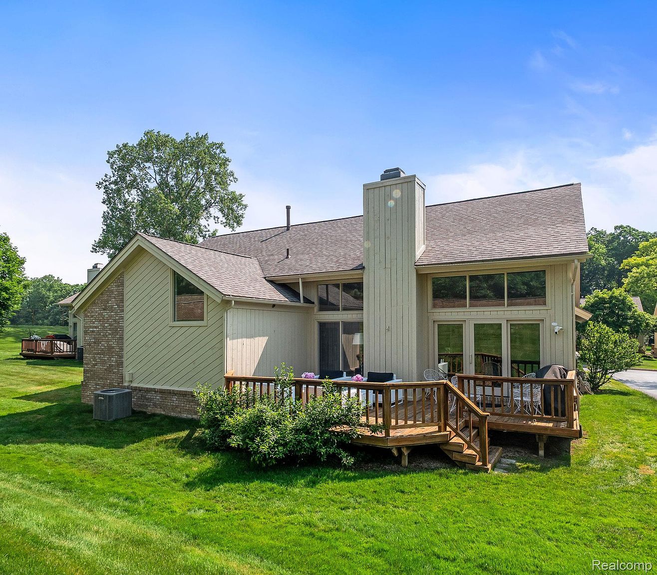 This is a rear view of a well-maintained house featuring a wooden deck with outdoor furniture, perfect for entertaining. The house has large windows offering plenty of natural light, and a brick chimney adds character. The lush green lawn and surrounding trees create a serene and private backyard setting.
