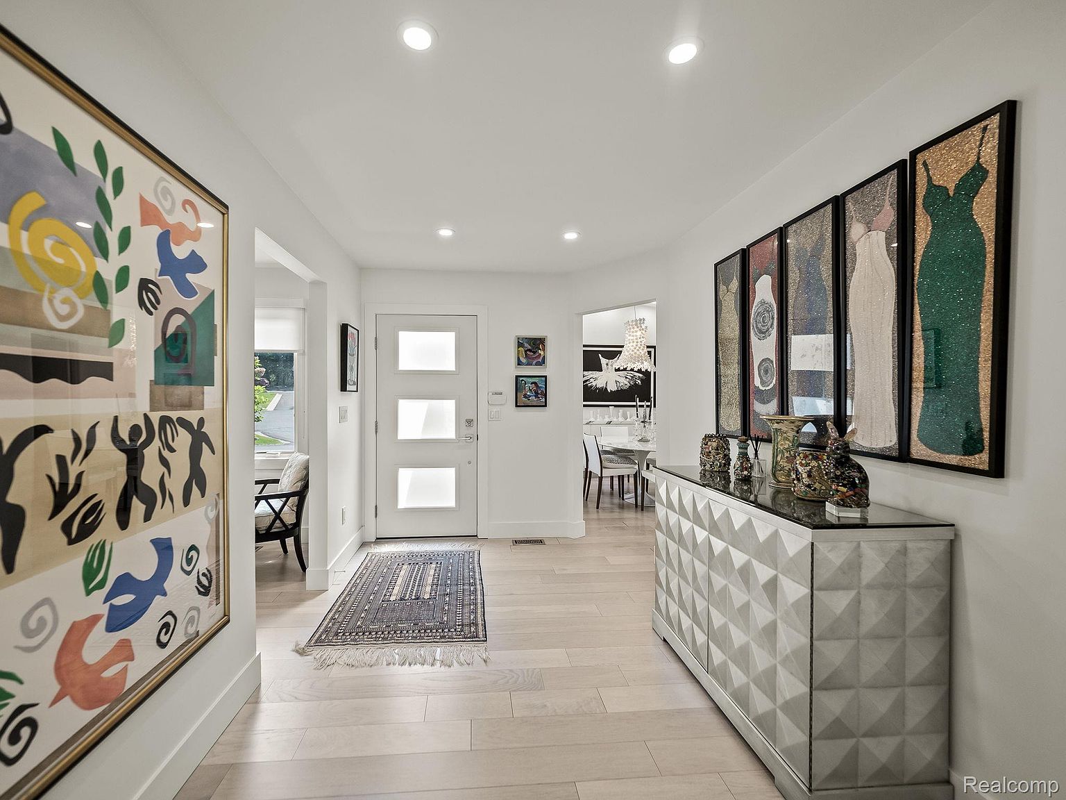 This is an interior shot of a bright and modern hallway. The walls are painted white, and the floors are light wood. The hallway features a large colorful painting on one side and a decorative cabinet with framed artwork above it on the other. A front door with frosted glass panels is visible at the end of the hallway, leading to the outside.