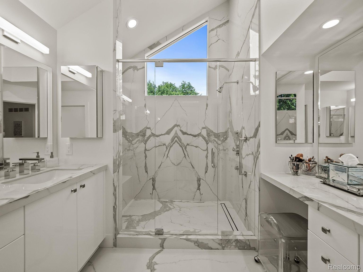 This is a bright and modern primary bathroom featuring a glass-enclosed shower with marble-patterned walls and a triangular window above. The bathroom includes a vanity with a white countertop and cabinetry, complemented by a large mirror and a separate makeup area with a clear acrylic chair. The overall design is clean, luxurious, and well-lit, creating a spa-like atmosphere.