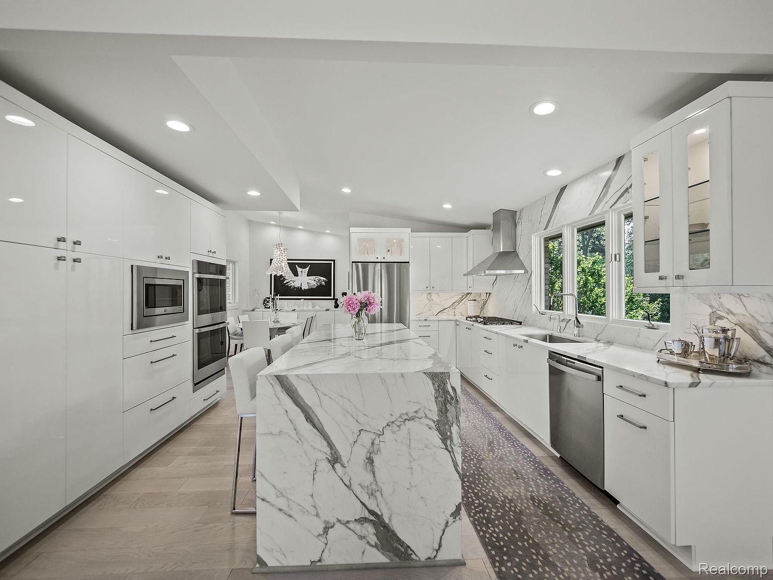 This is a bright, modern kitchen featuring sleek white cabinetry, stainless steel appliances, and a large marble island. The kitchen is well-lit with recessed lighting and natural light from the windows, which also provide a view of the outdoors. The perspective is a wide shot, showcasing the entire kitchen layout and design.