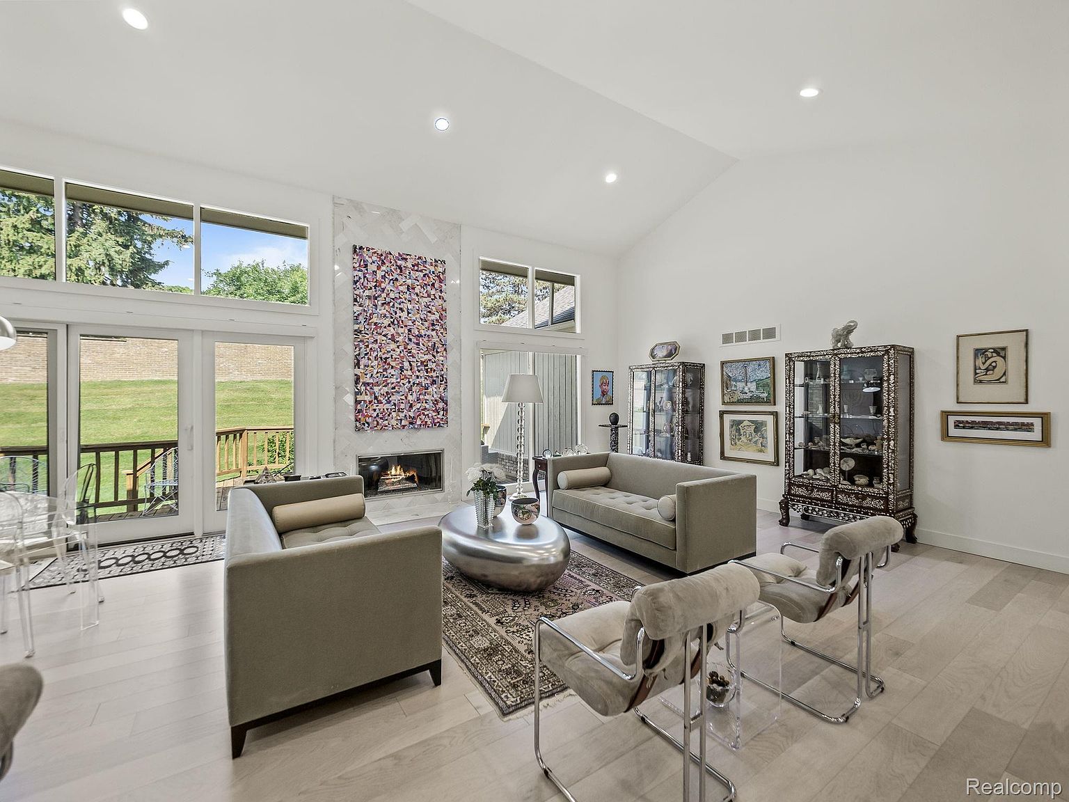 This is a bright and airy living room with a high vaulted ceiling and large windows offering natural light. The room features two sofas, a modern fireplace with a decorative marble surround, and unique display cabinets. The light wood flooring and neutral color palette create a sophisticated and inviting atmosphere.