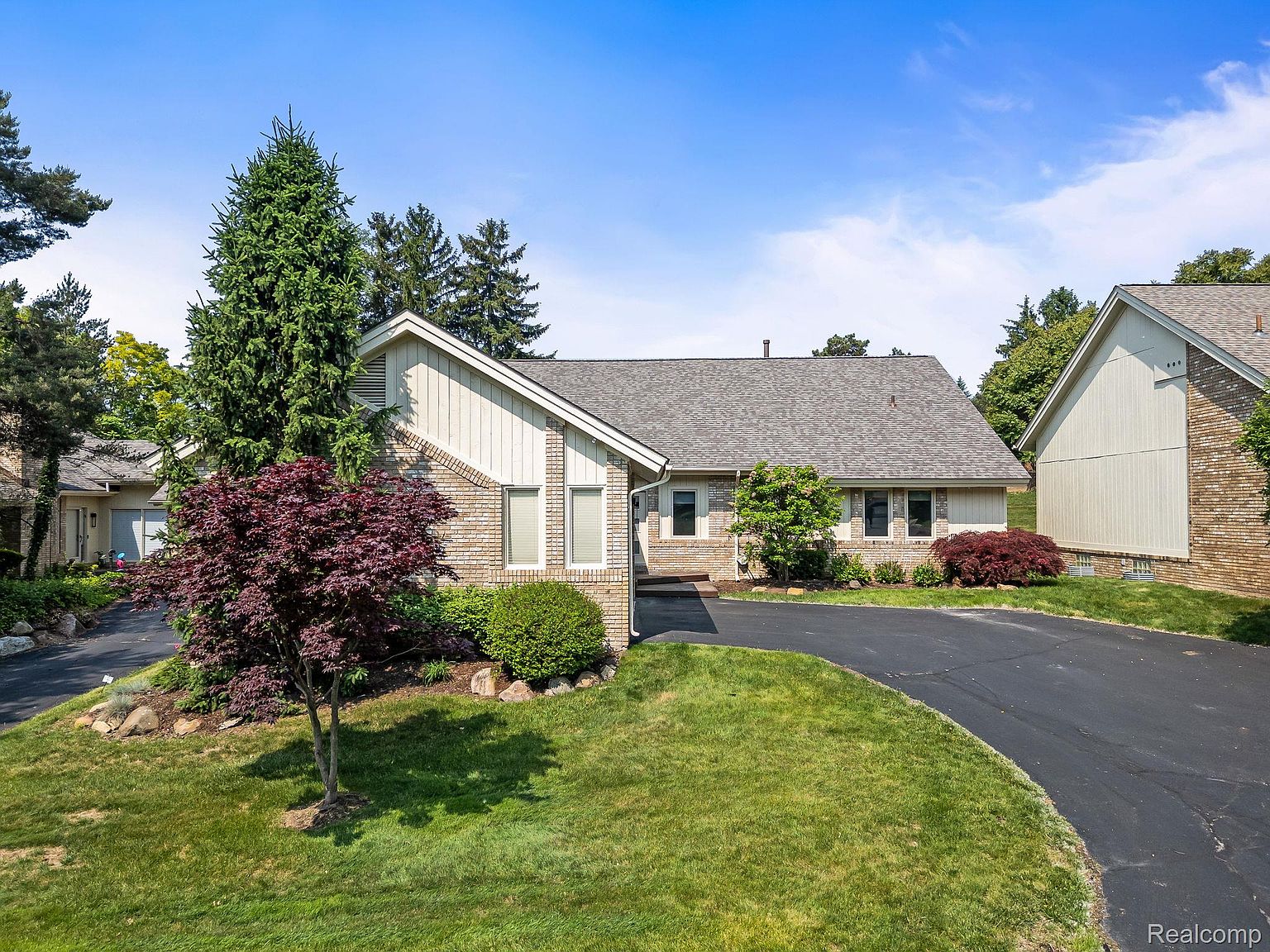 This is a front exterior view of a single-story brick and siding home with a well-manicured lawn and mature landscaping. A curved asphalt driveway leads to the house, enhancing its curb appeal. The roof is gray, and the home features a mix of brick and light-colored siding, giving it a classic and inviting appearance.