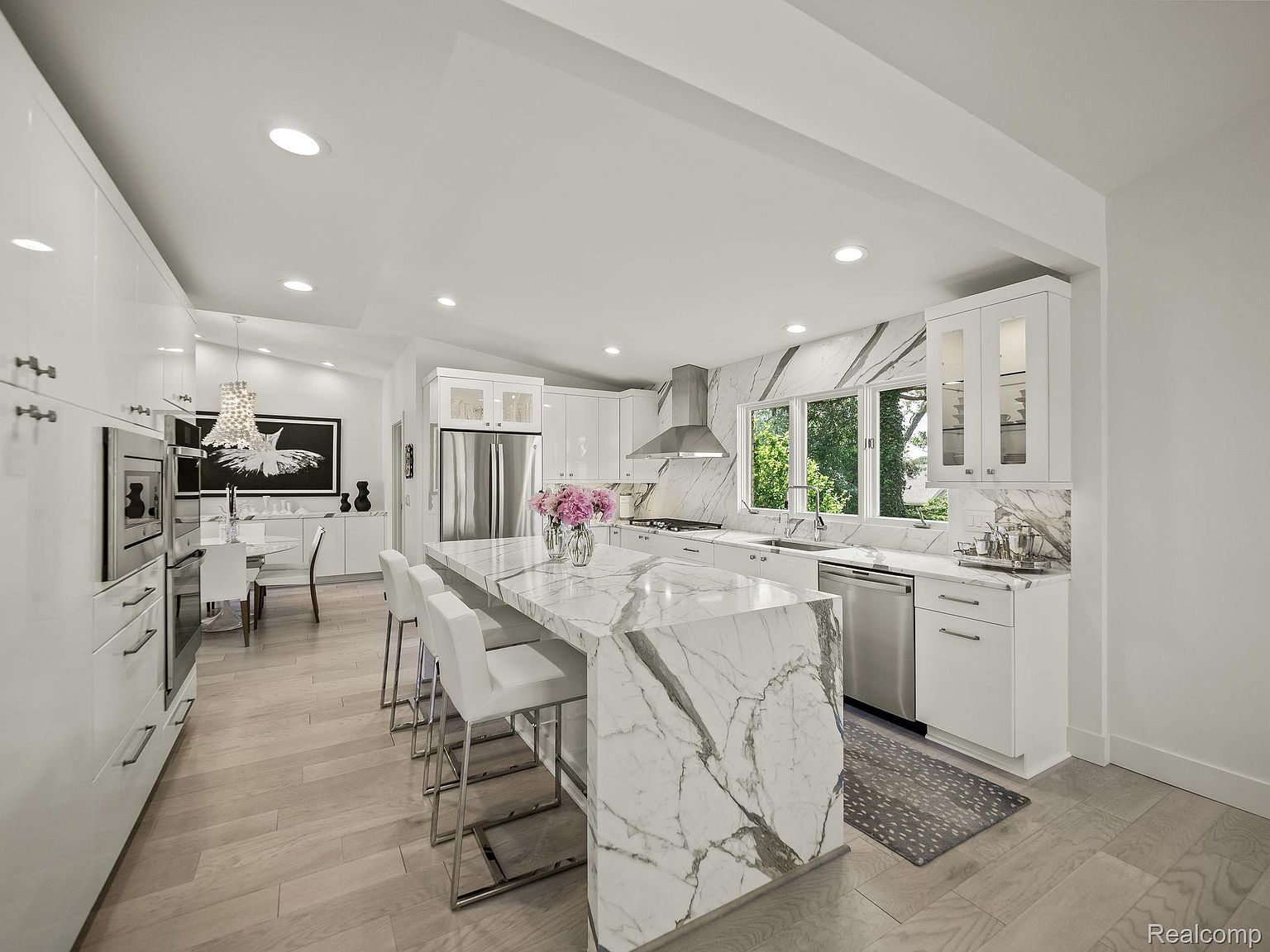 This is a bright and modern kitchen featuring white cabinetry, stainless steel appliances, and a large marble island with seating. The kitchen has a clean and contemporary style, with a large window providing natural light and a view of the outdoors. The perspective is a wide shot, showcasing the open layout and the connection to the dining area.