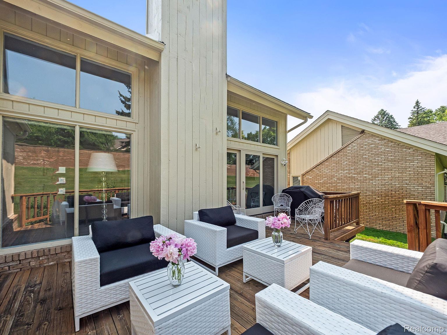 This is an exterior shot of a well-appointed patio or deck area. The space features modern white wicker furniture with black cushions, arranged around matching white tables adorned with floral arrangements. The deck is made of wood and is adjacent to a building with large windows and a brick exterior, suggesting a comfortable and stylish outdoor living space.