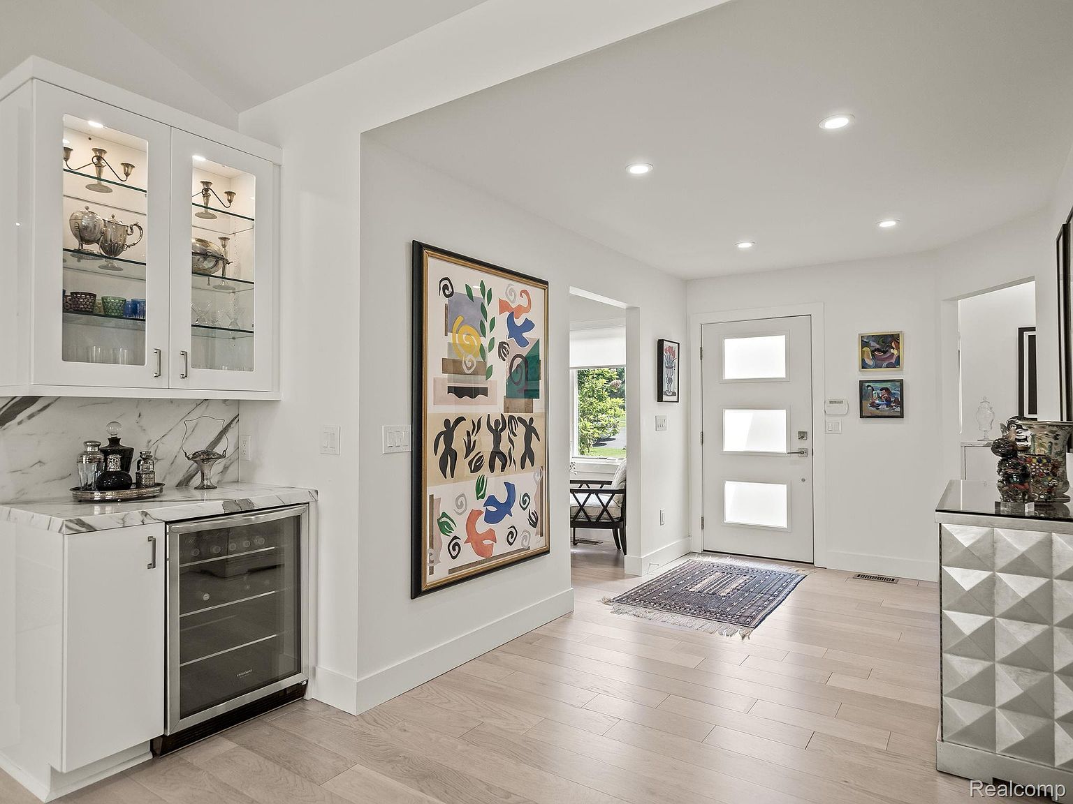 This interior shot showcases a bright and modern hallway with light wood flooring and white walls. A large, colorful abstract painting adds a focal point, while a built-in cabinet with a wine fridge provides a touch of luxury. The space is well-lit with recessed lighting and natural light filtering through the doorway, creating an inviting and airy atmosphere.