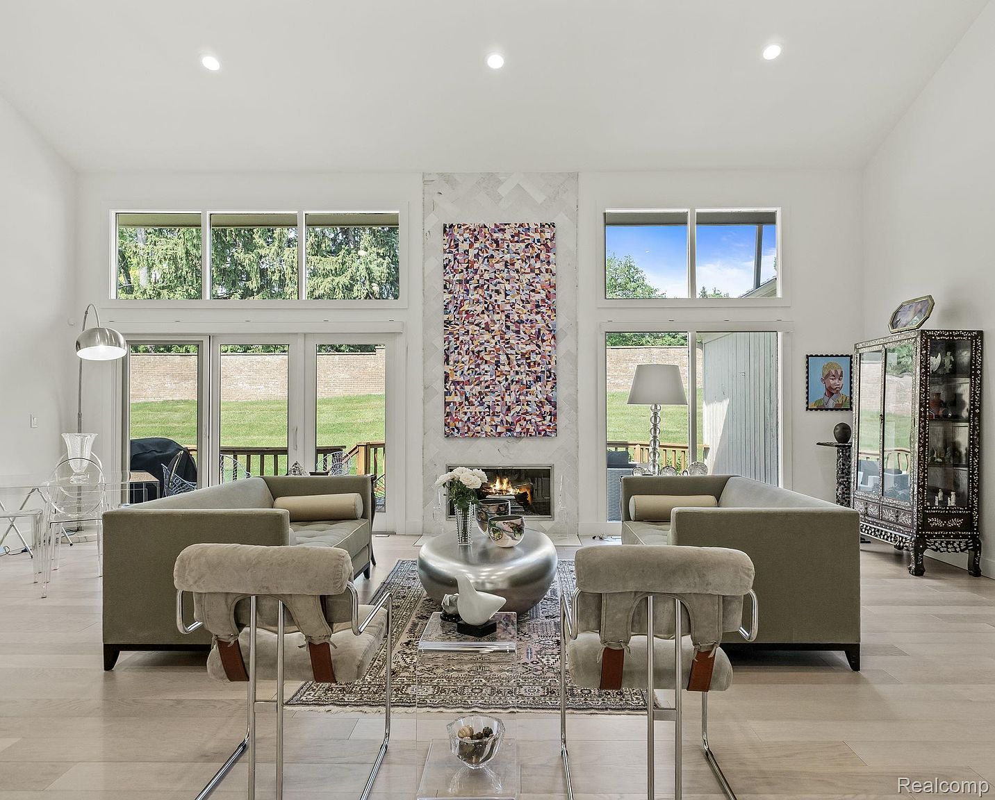 This is an interior shot of a modern living room featuring two sofas facing each other, a unique silver coffee table, and two stylish chairs in the foreground. A large abstract art piece hangs above the fireplace, flanked by large windows that offer views of the outdoors. The room is bright and airy, with light hardwood floors and a neutral color palette, creating a sophisticated and inviting atmosphere.