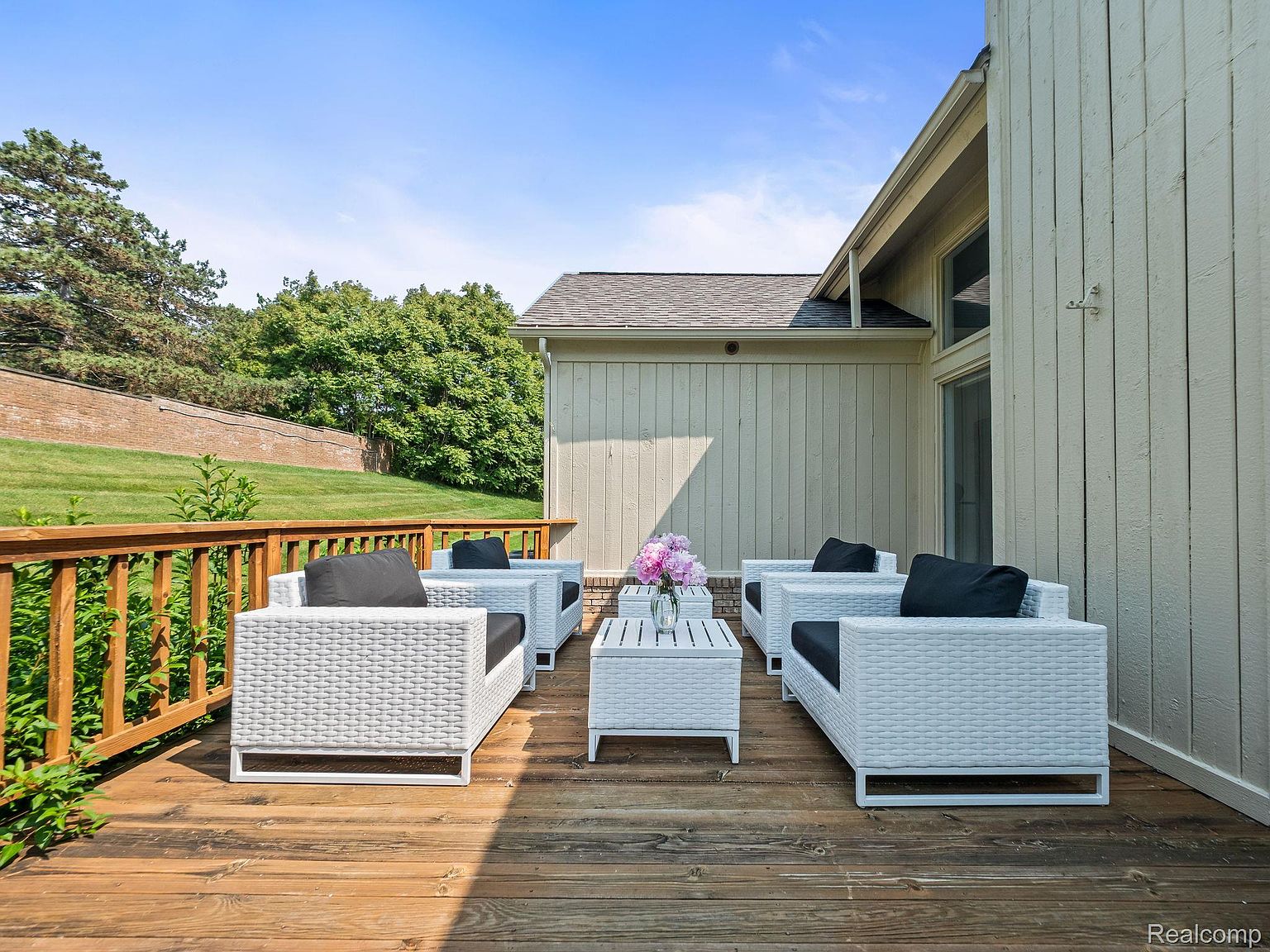 This image showcases a well-appointed outdoor patio or deck area, featuring modern white wicker furniture with black cushions arranged around a matching coffee table. The wooden deck provides a warm contrast to the furniture, and the surrounding landscape includes a manicured lawn, trees, and a partial view of the house's exterior, creating an inviting and stylish outdoor living space.